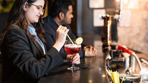 A person holding a cocktail glass with a lemon slice, seated at a bar with a stainless steel counter. - Novotel Milton Keynes (Milton Keynes)