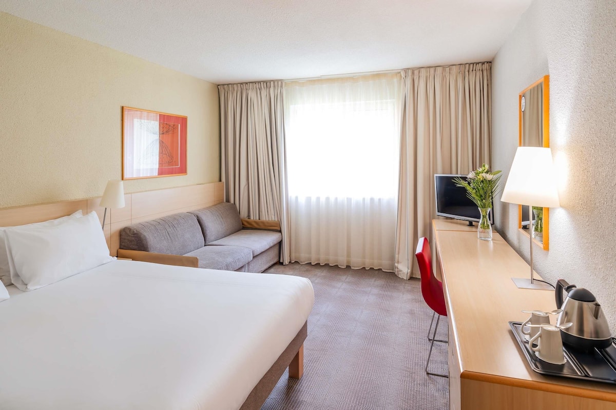 A hotel room with a bed, a sofa, a desk with a lamp, a TV, and a tray with a kettle and cups.
