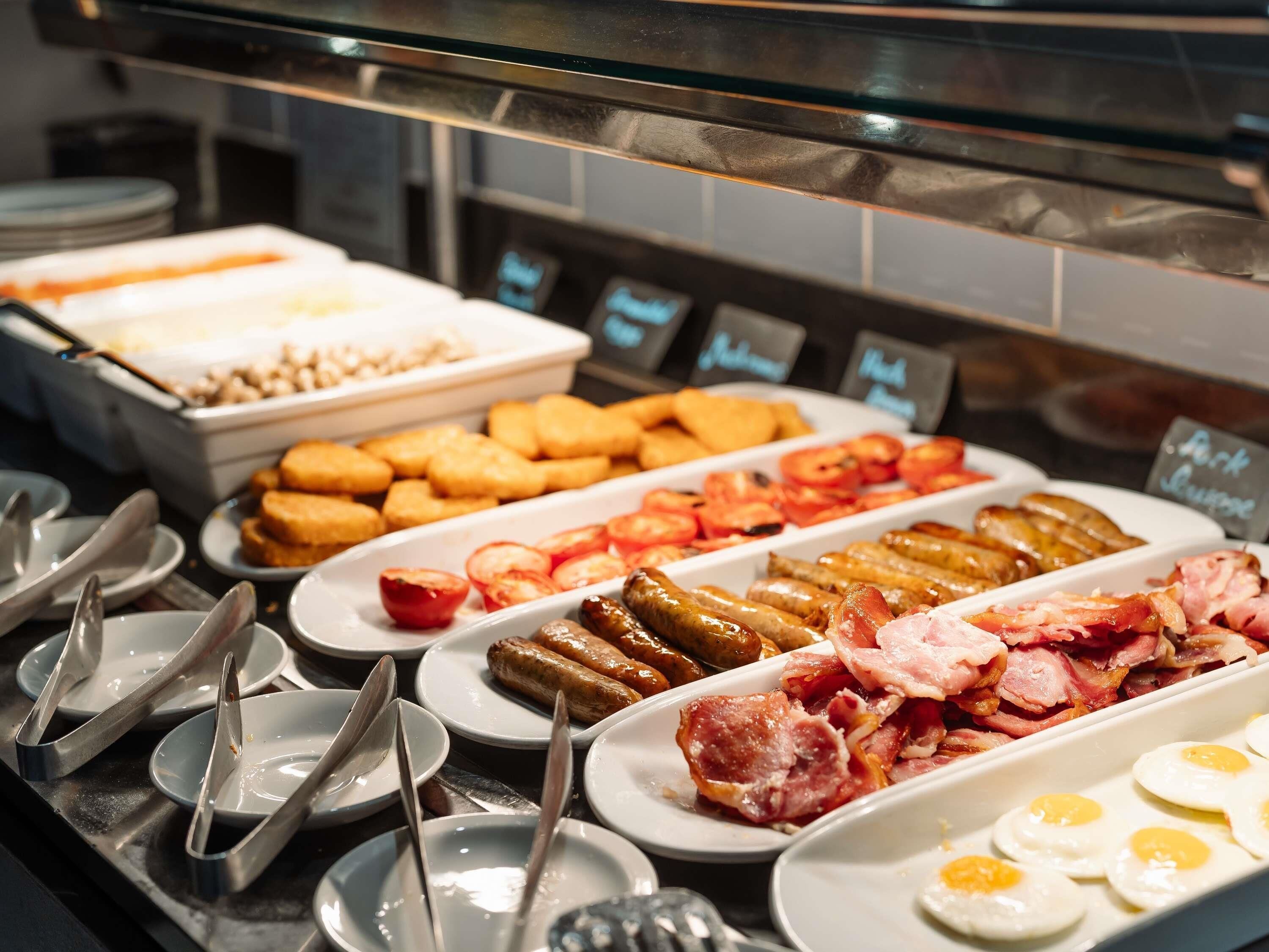 Een buffet met diverse etenswaren, waaronder worstjes, spek, eieren en plakjes tomaat.