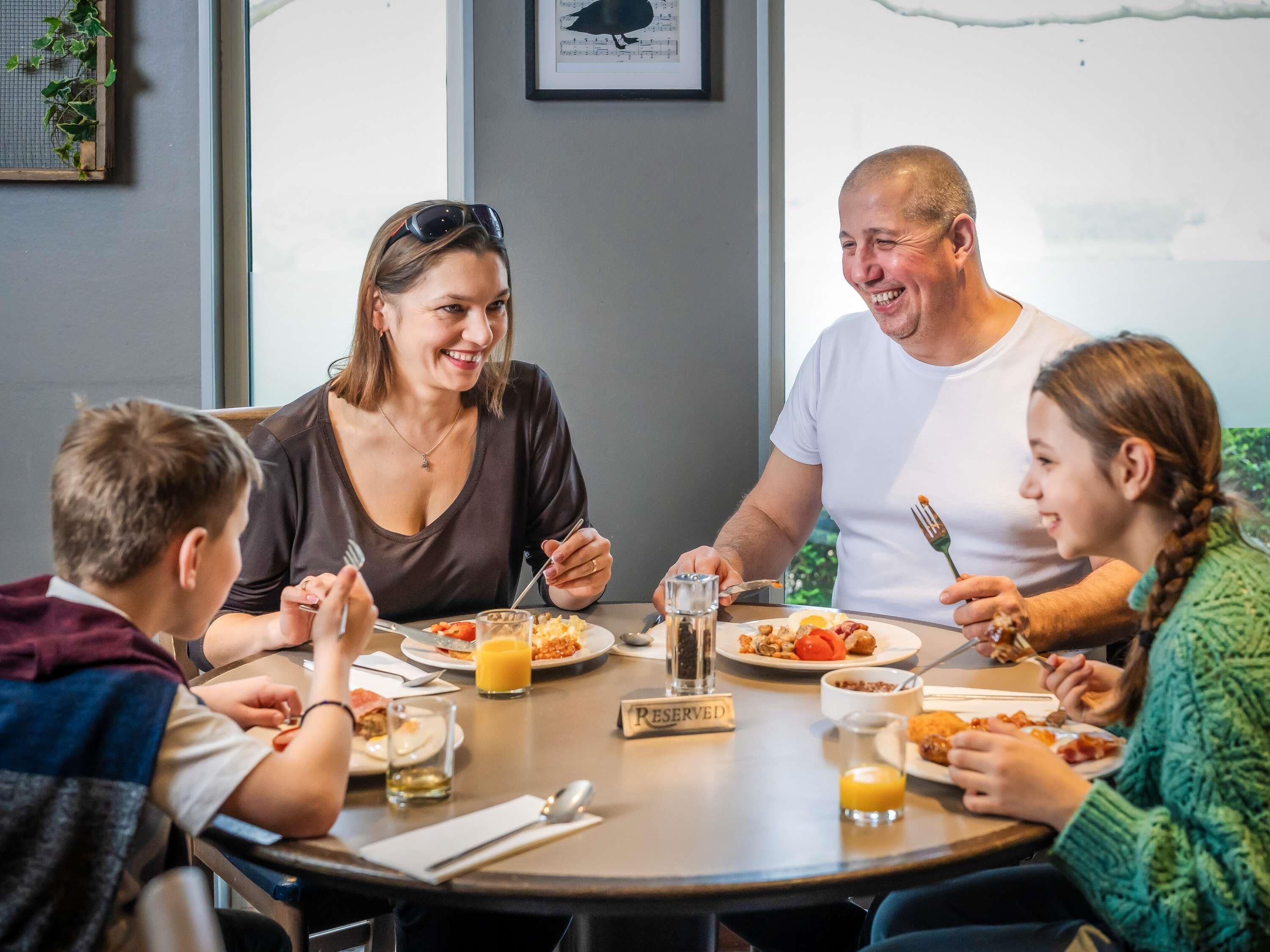 Une famille est assise autour d’une table ronde, où l’on peut voir différents plats et boissons. Une pancarte « RÉSERVÉ » est posée sur la table, et une fenêtre offre une vue sur l’extérieur.