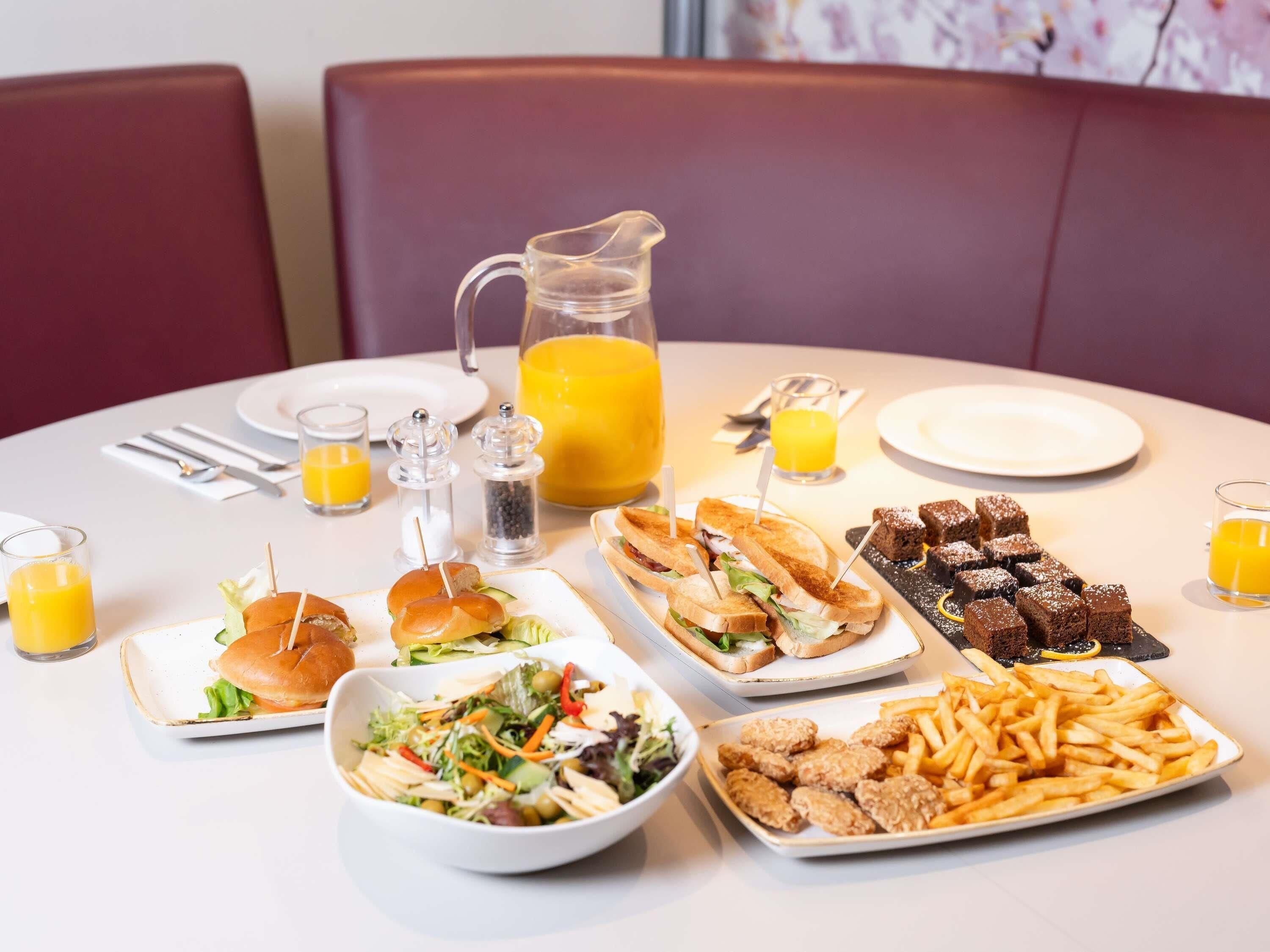 Une table à manger avec divers aliments pour le petit-déjeuner, notamment des sandwichs, une carafe de jus d’orange et une salade.