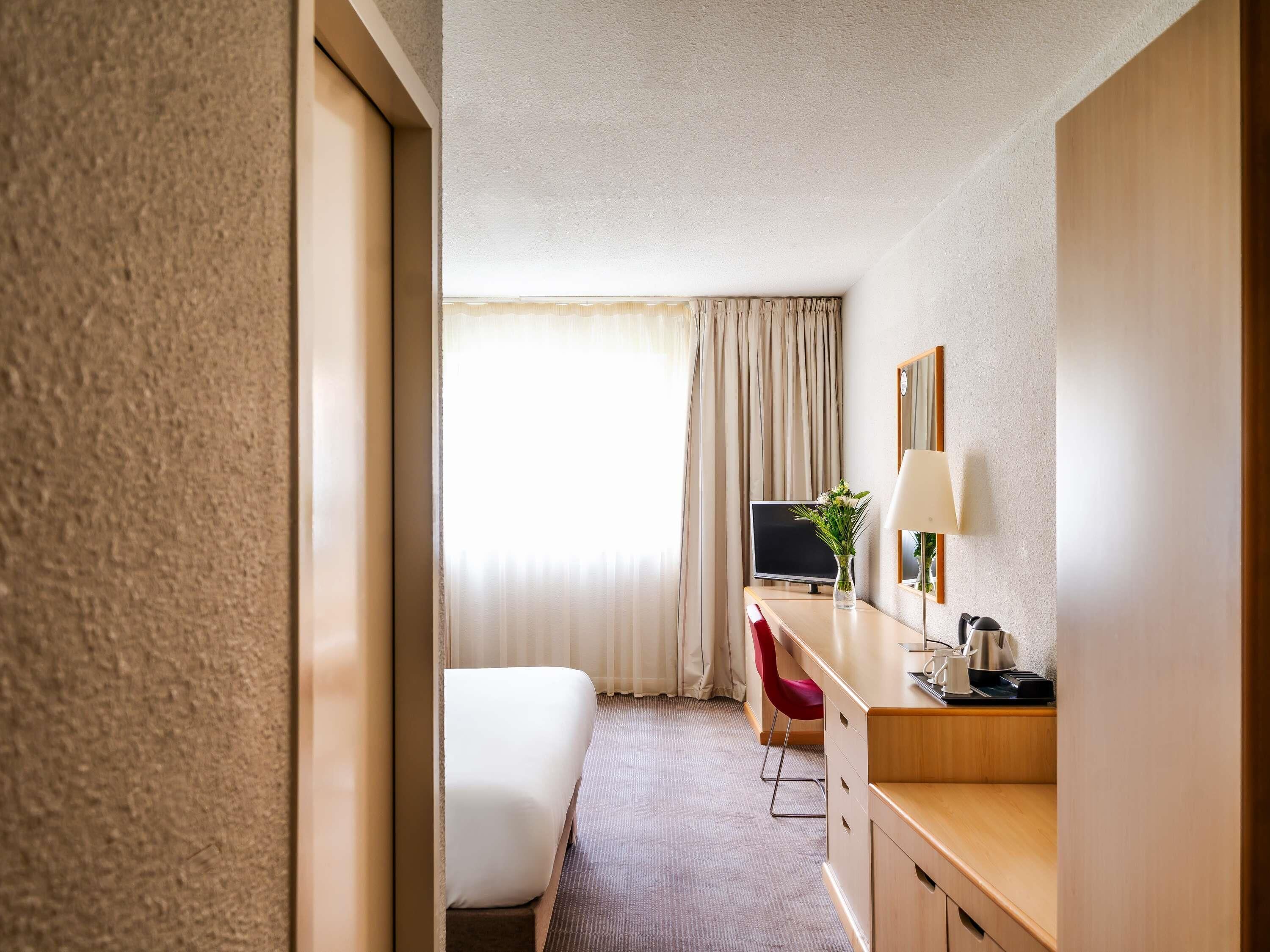 A hotel room with a bed, a desk with a TV, a chair, a lamp, and a mirror.