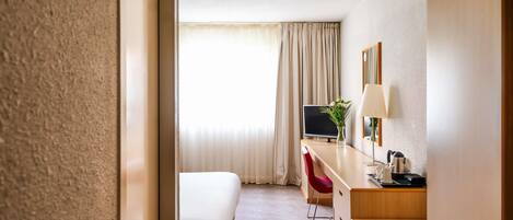 A hotel room with a bed, a desk with a TV, a chair, a lamp, and a mirror.