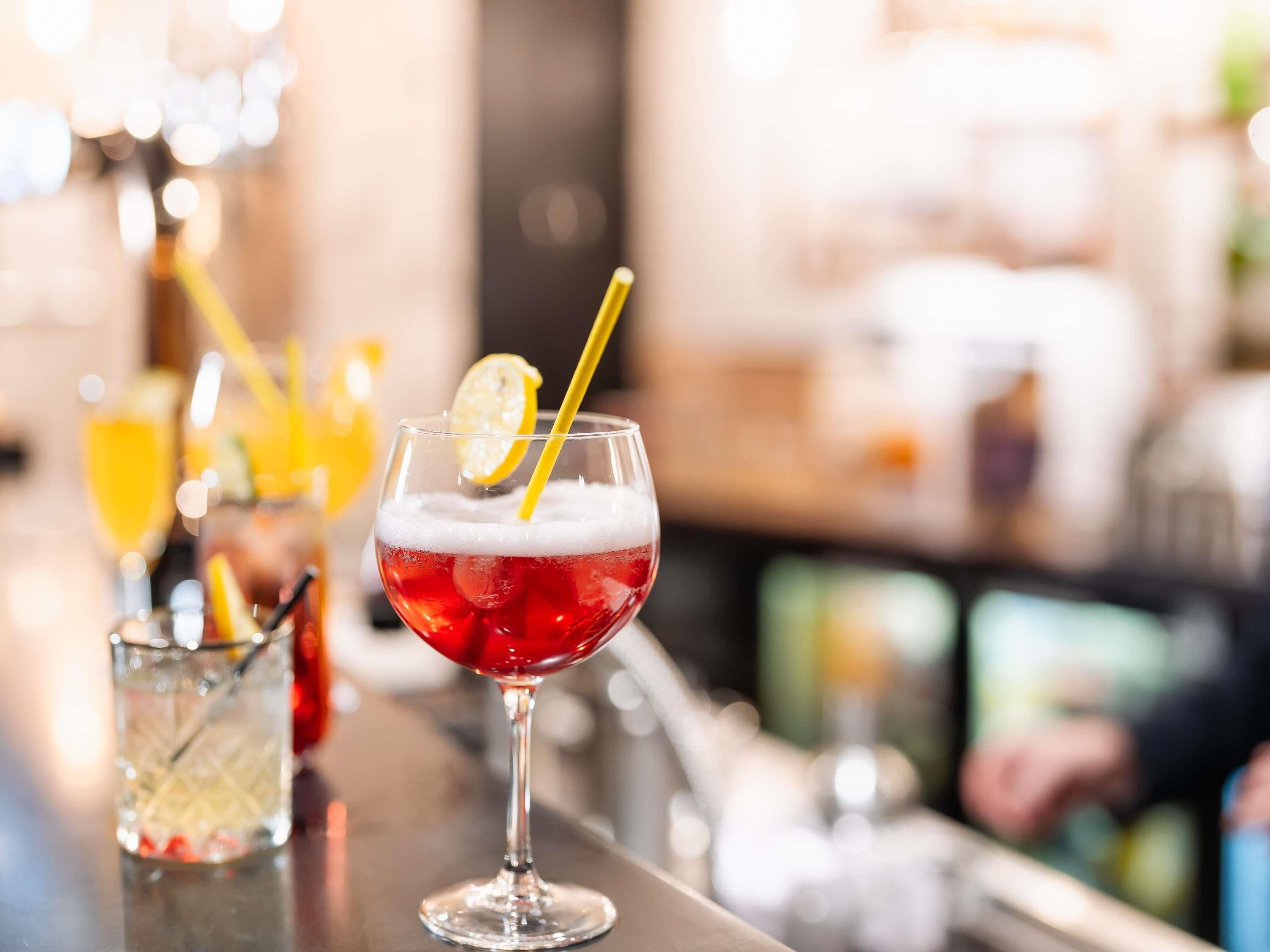 Un bancone bar con un bicchiere di cocktail rosso con ghiaccio, una fetta di limone e una cannuccia.