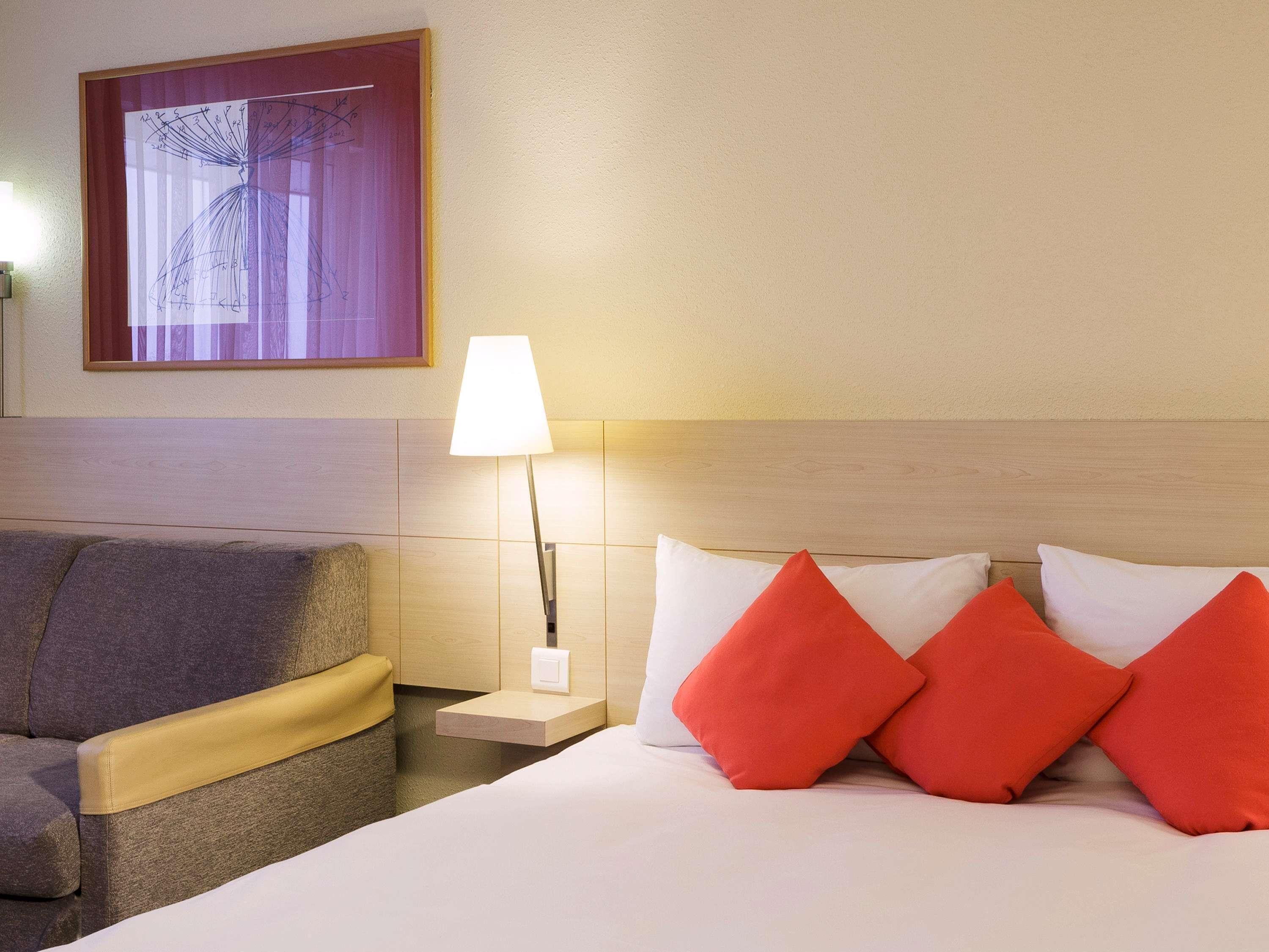 A hotel room with a bed, a nightstand, a lamp, a couch, and a window with curtains.