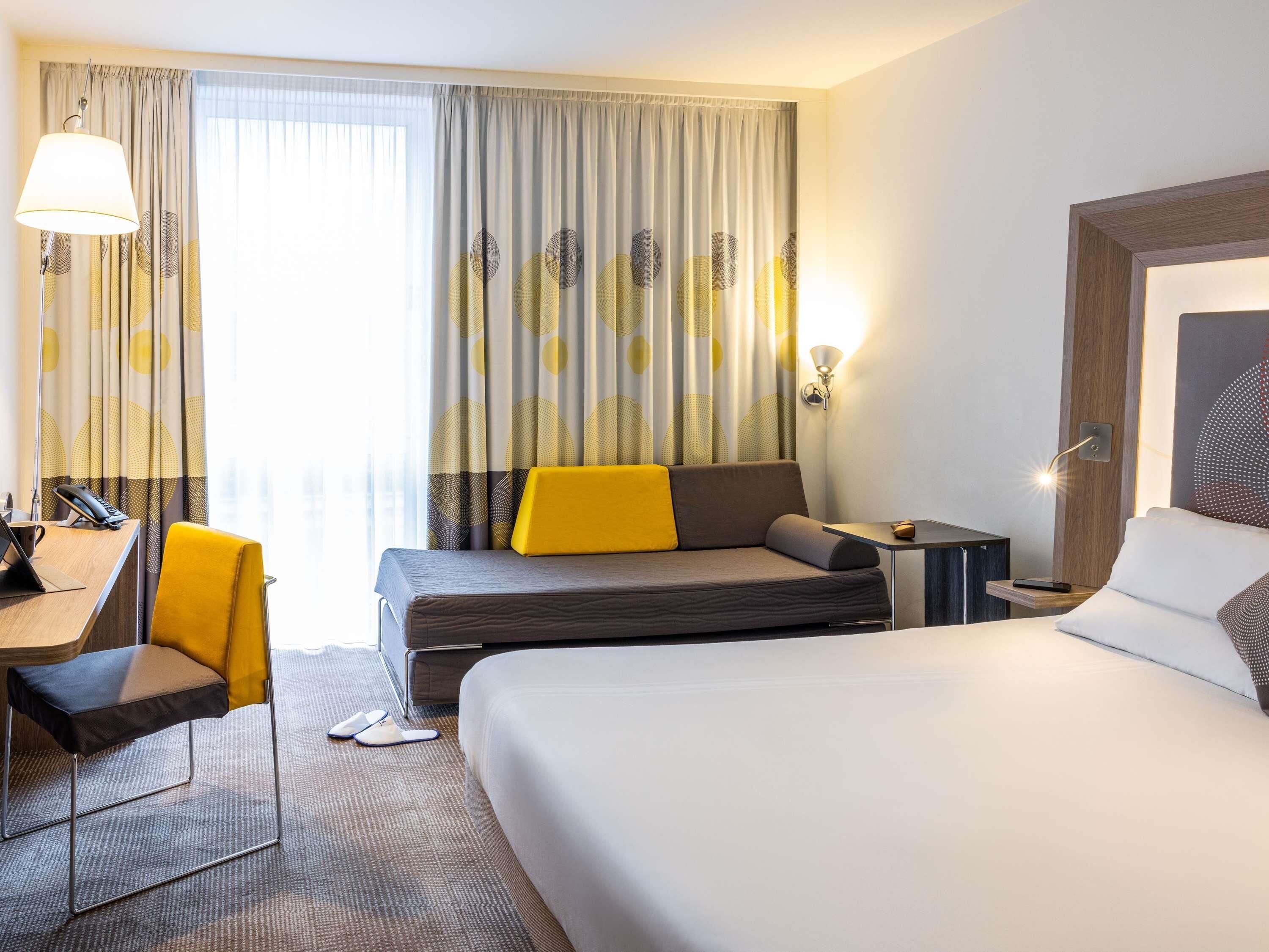 A hotel room with a bed, a desk, a chair, a sofa, and a large window with curtains.