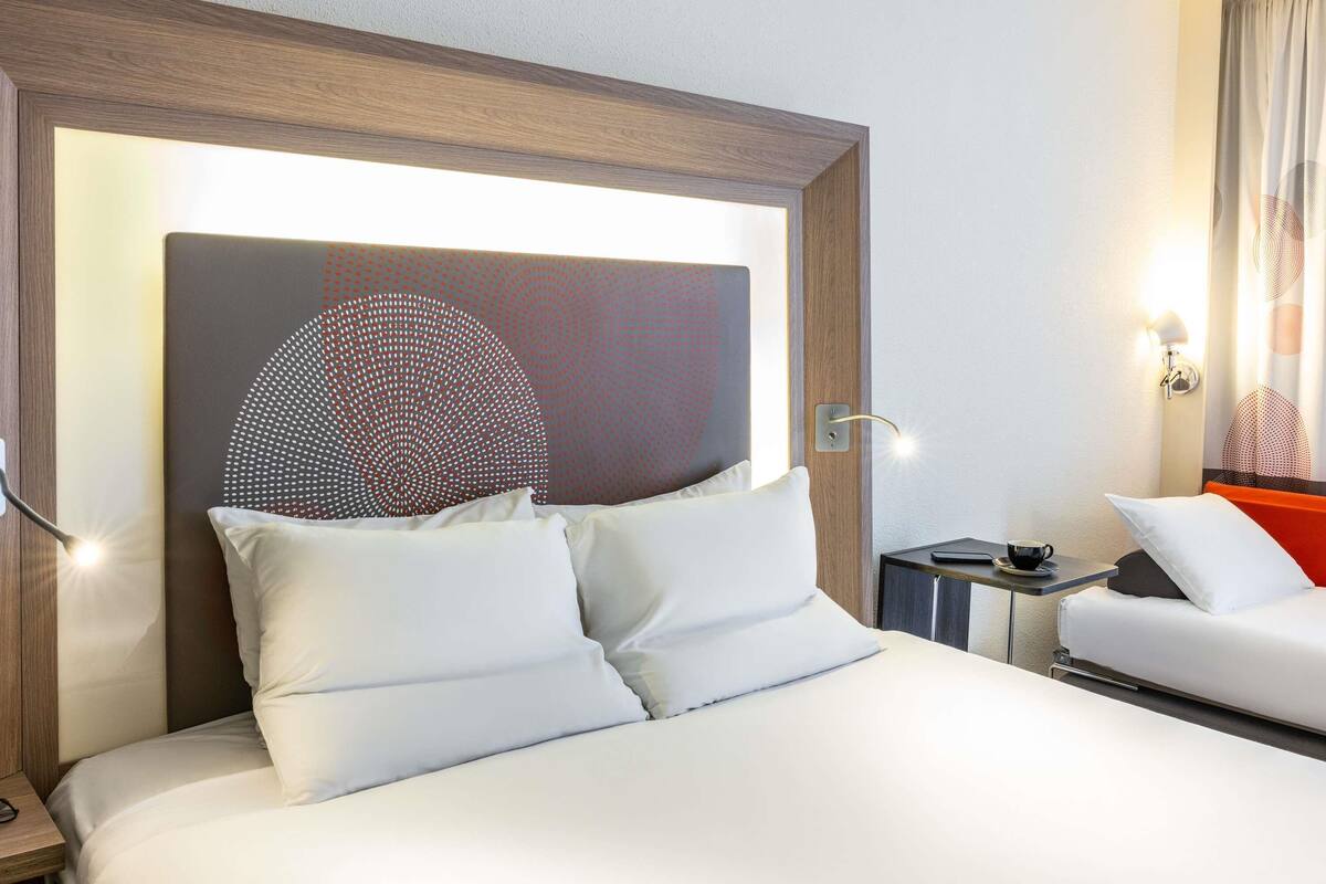 A hotel room with a bed, a nightstand, a chair, and a wall-mounted lamp.
