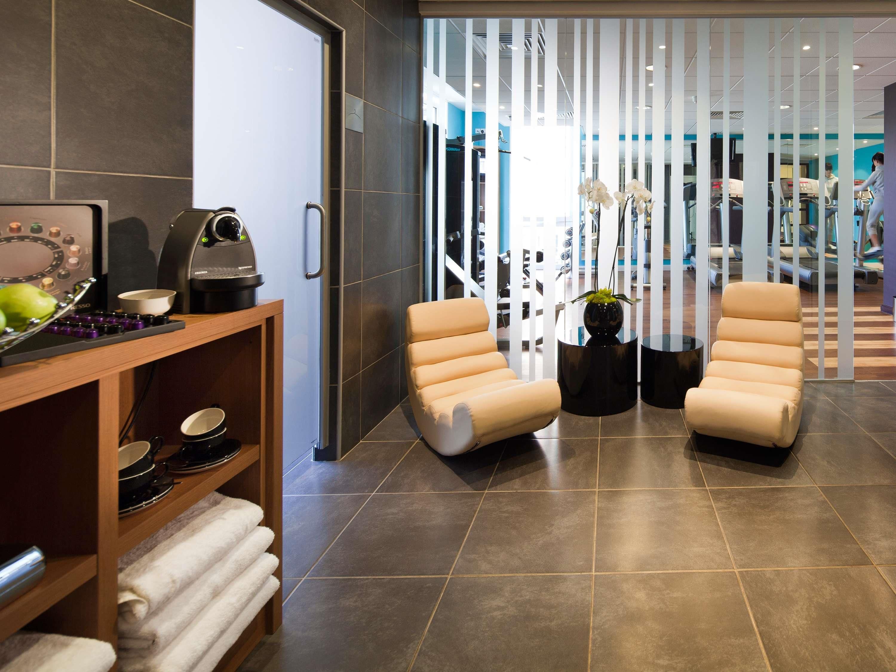 A modern lobby with a reception desk, two leather armchairs, a coffee machine, and a potted plant.