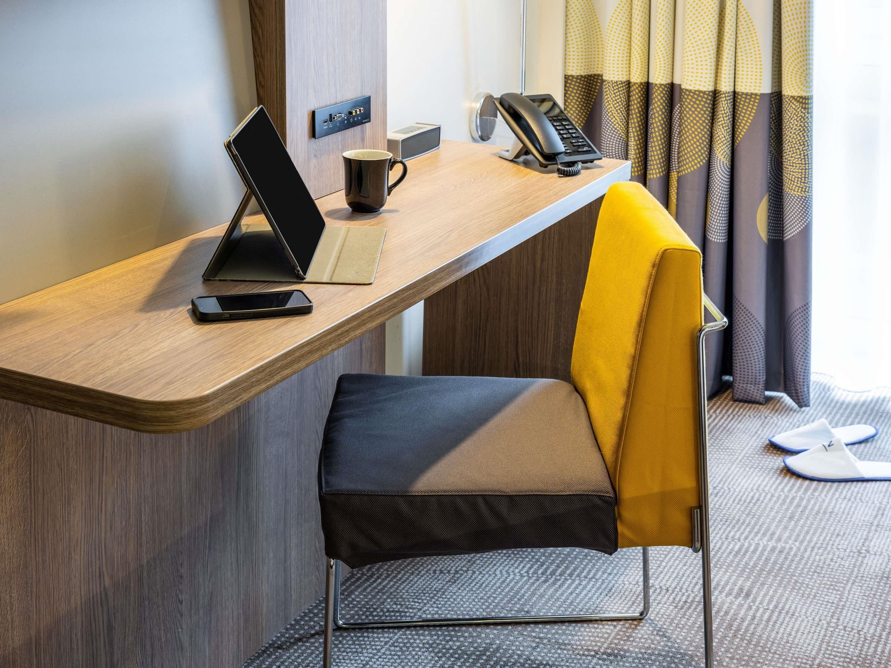 A hotel room with a desk, a laptop, a mobile phone, a telephone, a cup, and a chair.