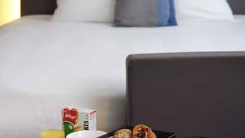 Una bandeja de desayuno ordenada con diversos alimentos y una jarra de agua, colocada frente a una cama con cabecera y almohadas.