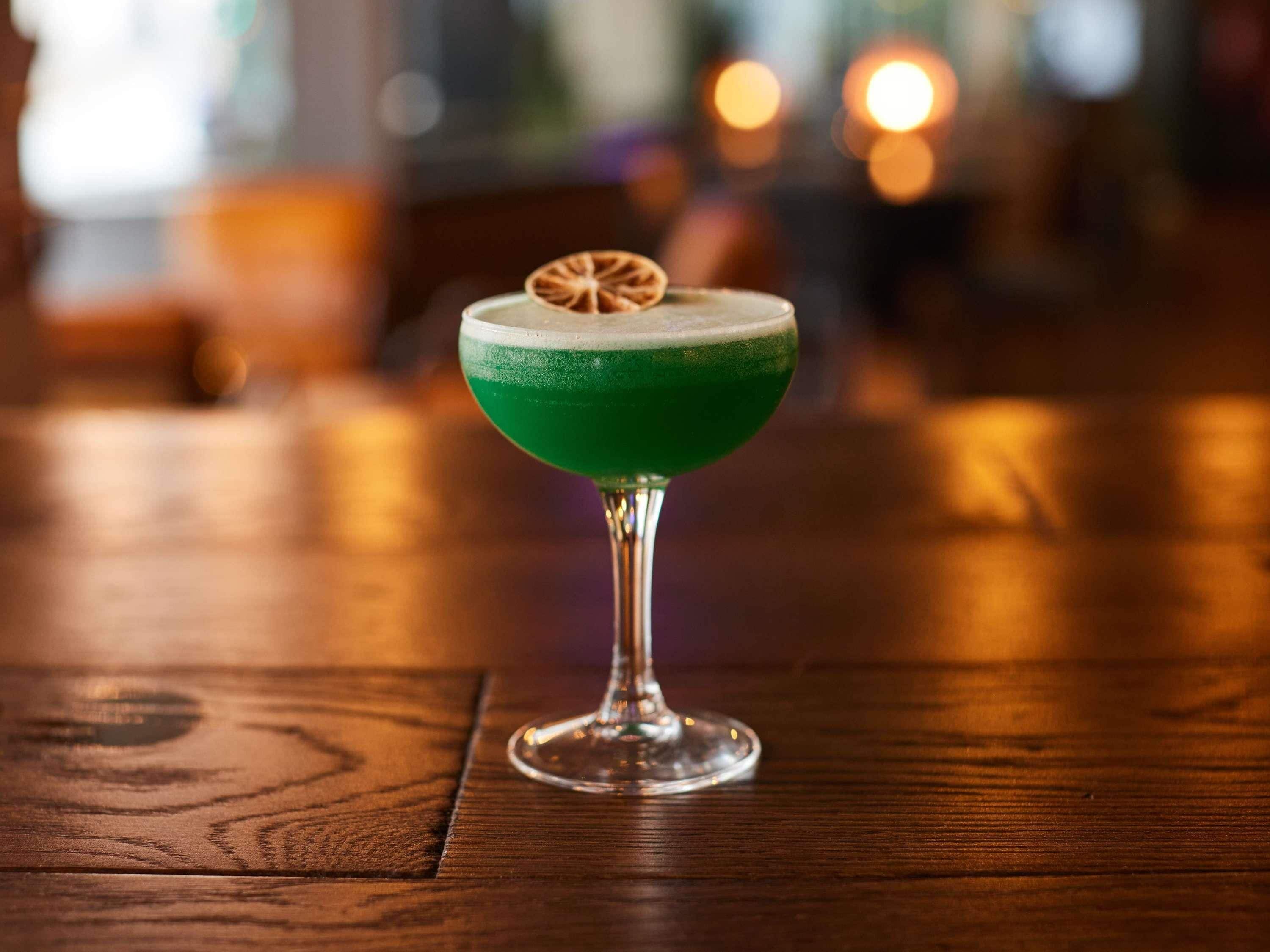A green cocktail in a stemmed glass with a dried orange slice garnish on a wooden table.