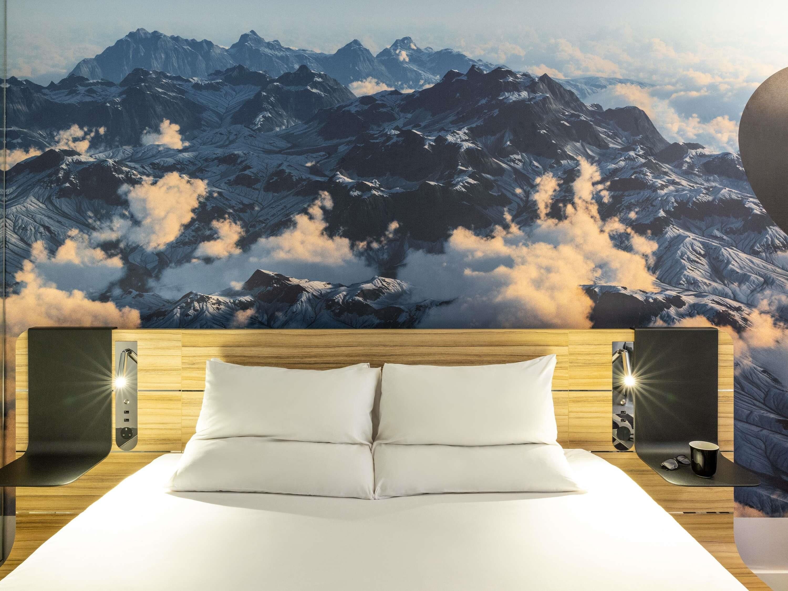 Ein Hotelzimmer mit einem Bett, einem Holzkopfteil, zwei Nachttischen, einem Wandbild von Bergen und einem Beistelltisch mit einer Lampe.