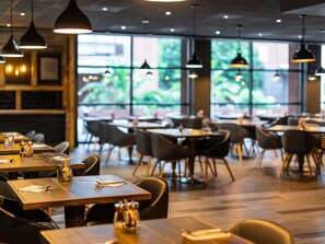 A modern restaurant interior with wooden tables, black chairs, pendant lighting, and large windows with a view of greenery. - Novotel Leeds Centre (Leeds)