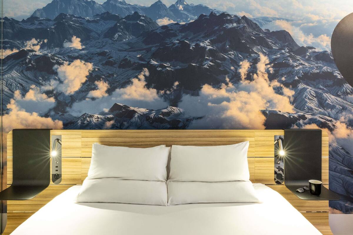 A hotel room with a bed, wooden headboard, two bedside tables, a wall mural of mountains, and a nightstand with a lamp.