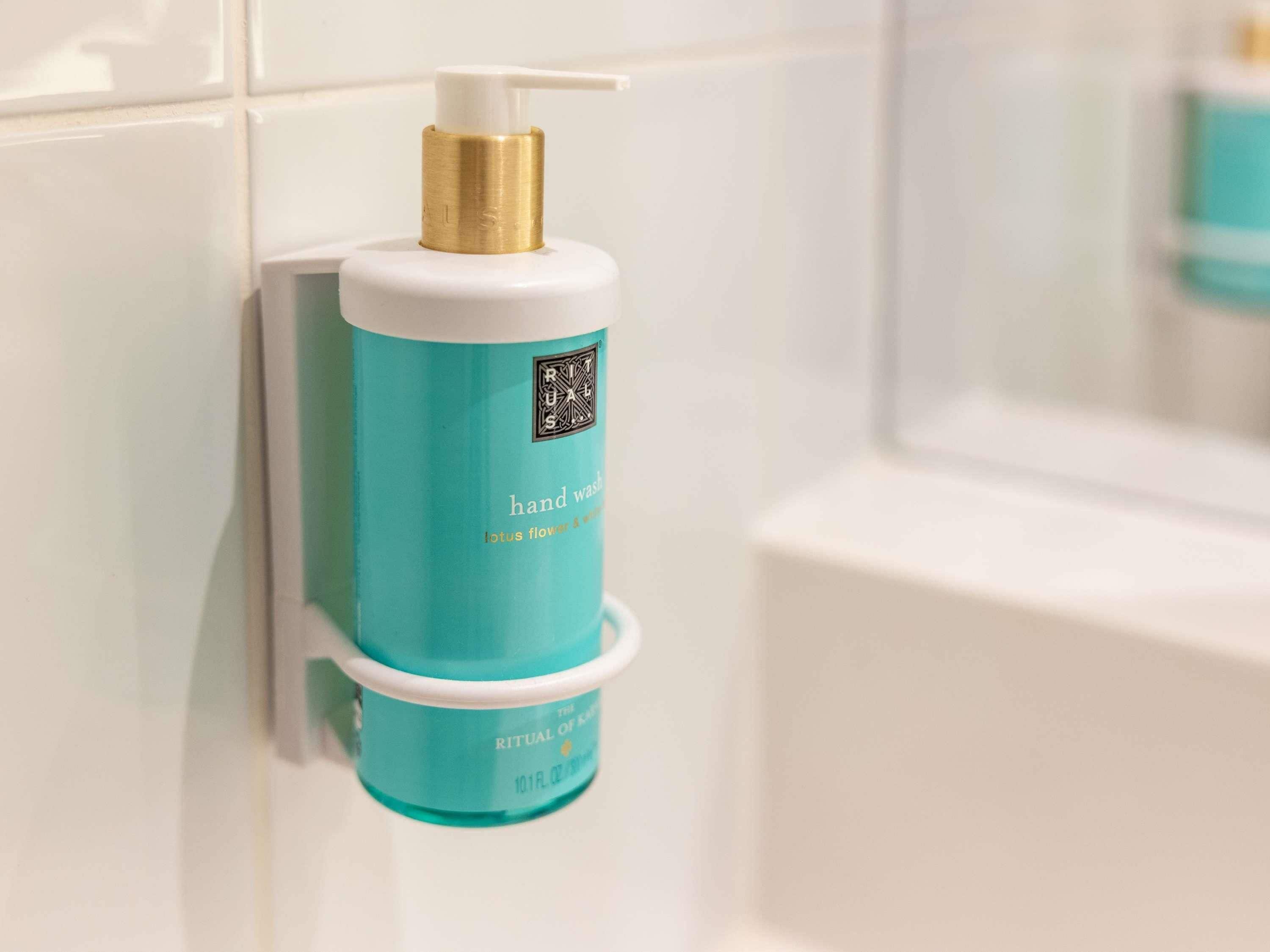 A hand wash bottle with a pump, labeled 'RITUAL OF KAR' and 'hand wash lotus flower & rose,' mounted on a white tiled wall.