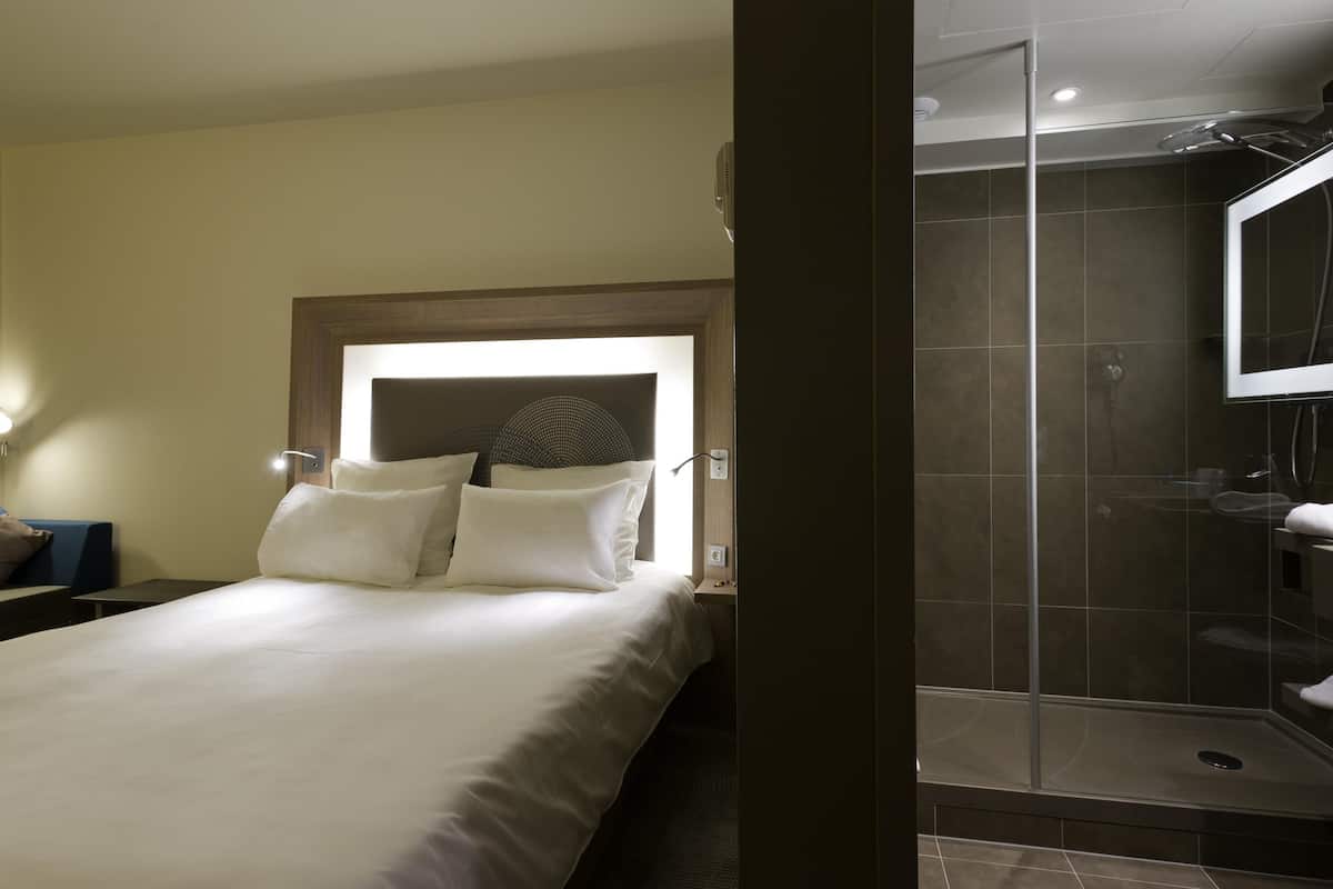 A hotel room with a bed, bedside table, lamp, and a glass-enclosed shower area.