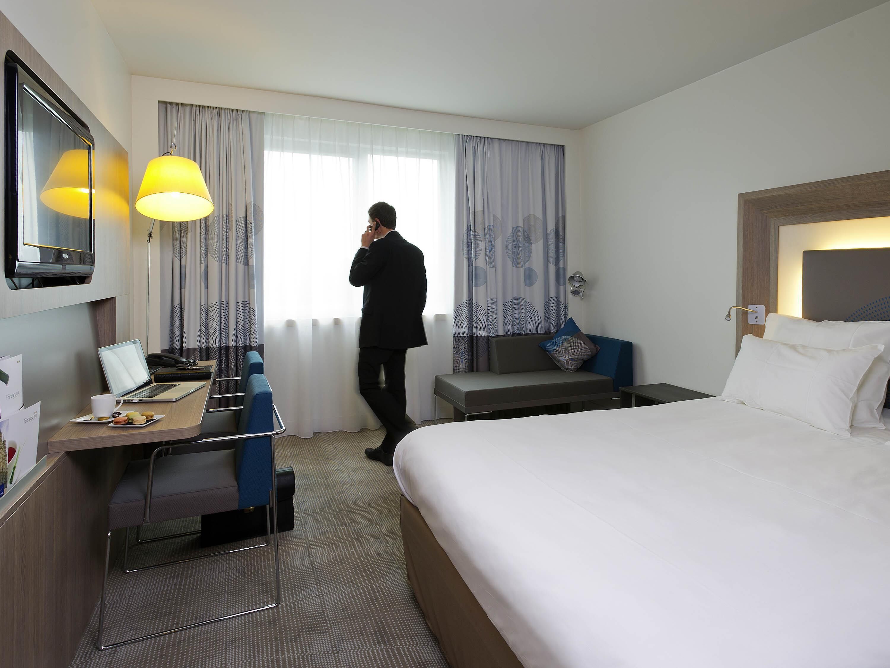 Een hotelkamer met een bed, een bureau met een laptop, een stoel, een bank en een groot raam met gordijnen.