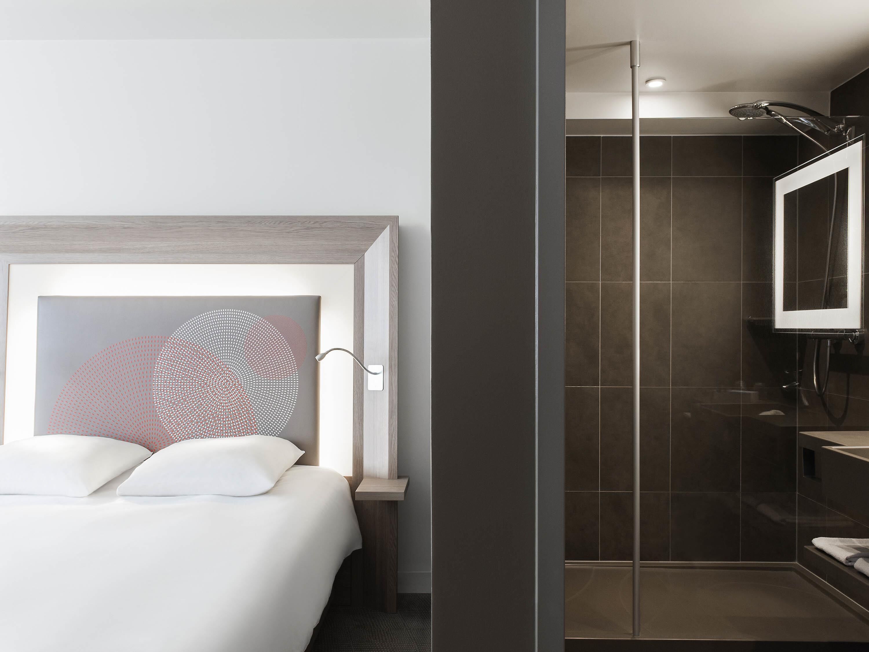 A modern hotel room with a bed, a shower area, and a large mirror.