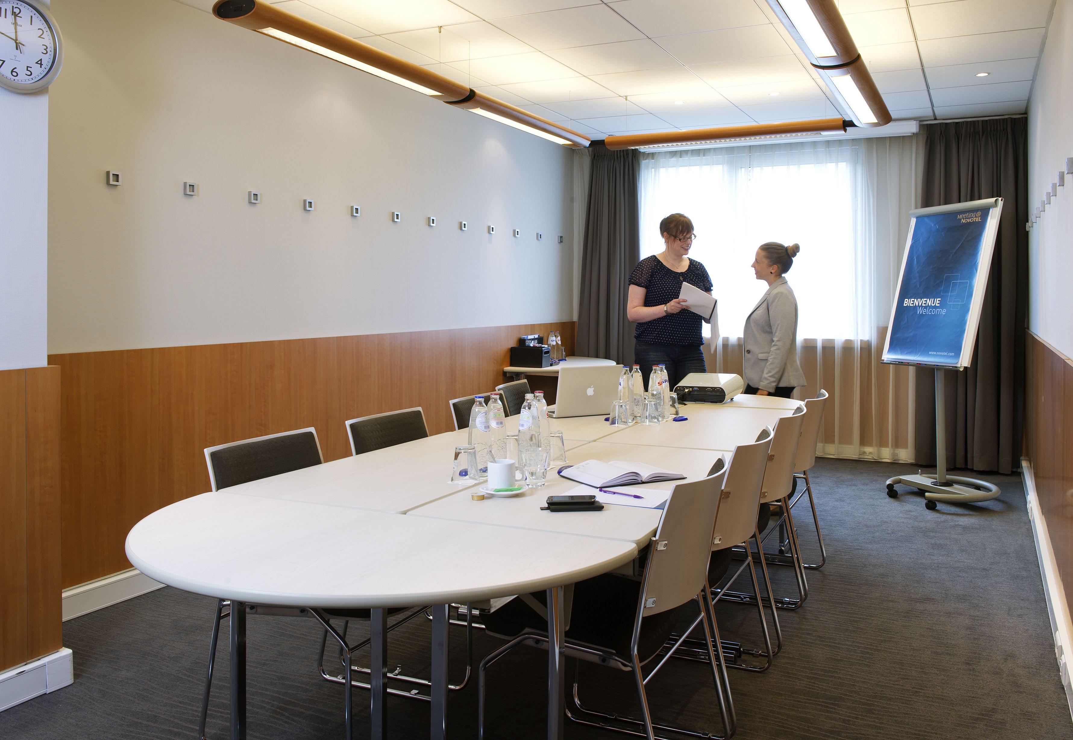 A conference room with a long table, chairs, a clock, and a welcome sign.