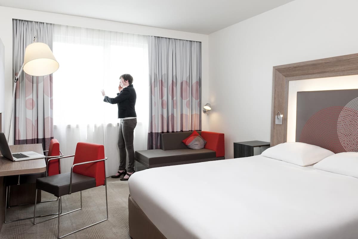 A hotel room with a bed, a desk with a laptop, a chair, a sofa, and a window with curtains.