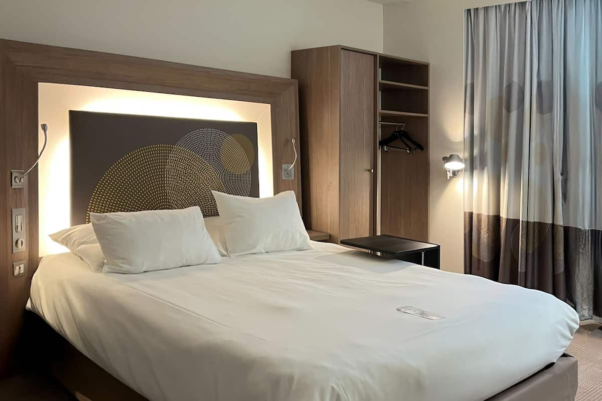 A hotel room with a large bed, a wooden headboard, a wardrobe, and a curtain.