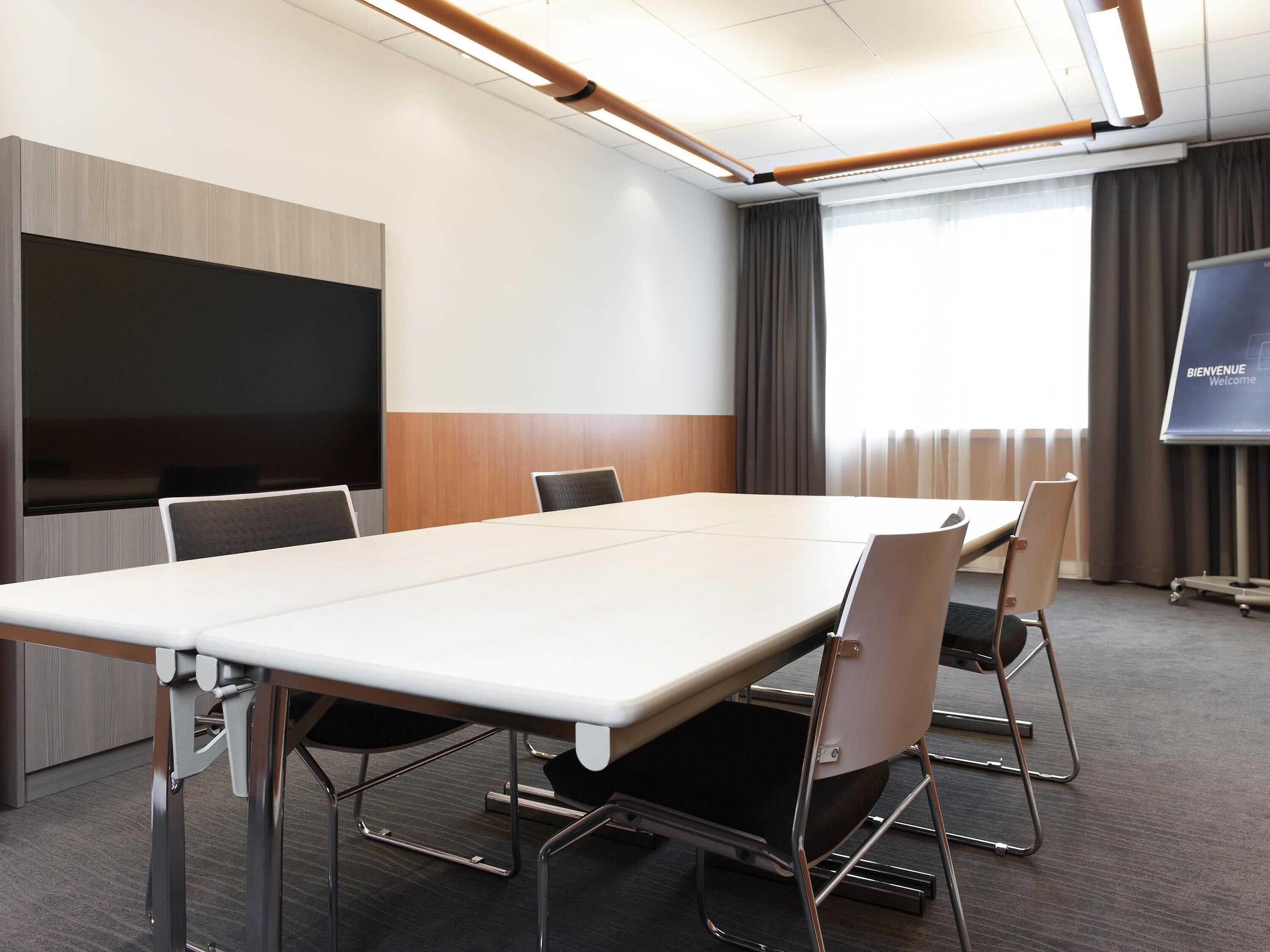 A conference room with a long table, chairs, a TV screen, and a 'BIENVENUE Welcome' sign.