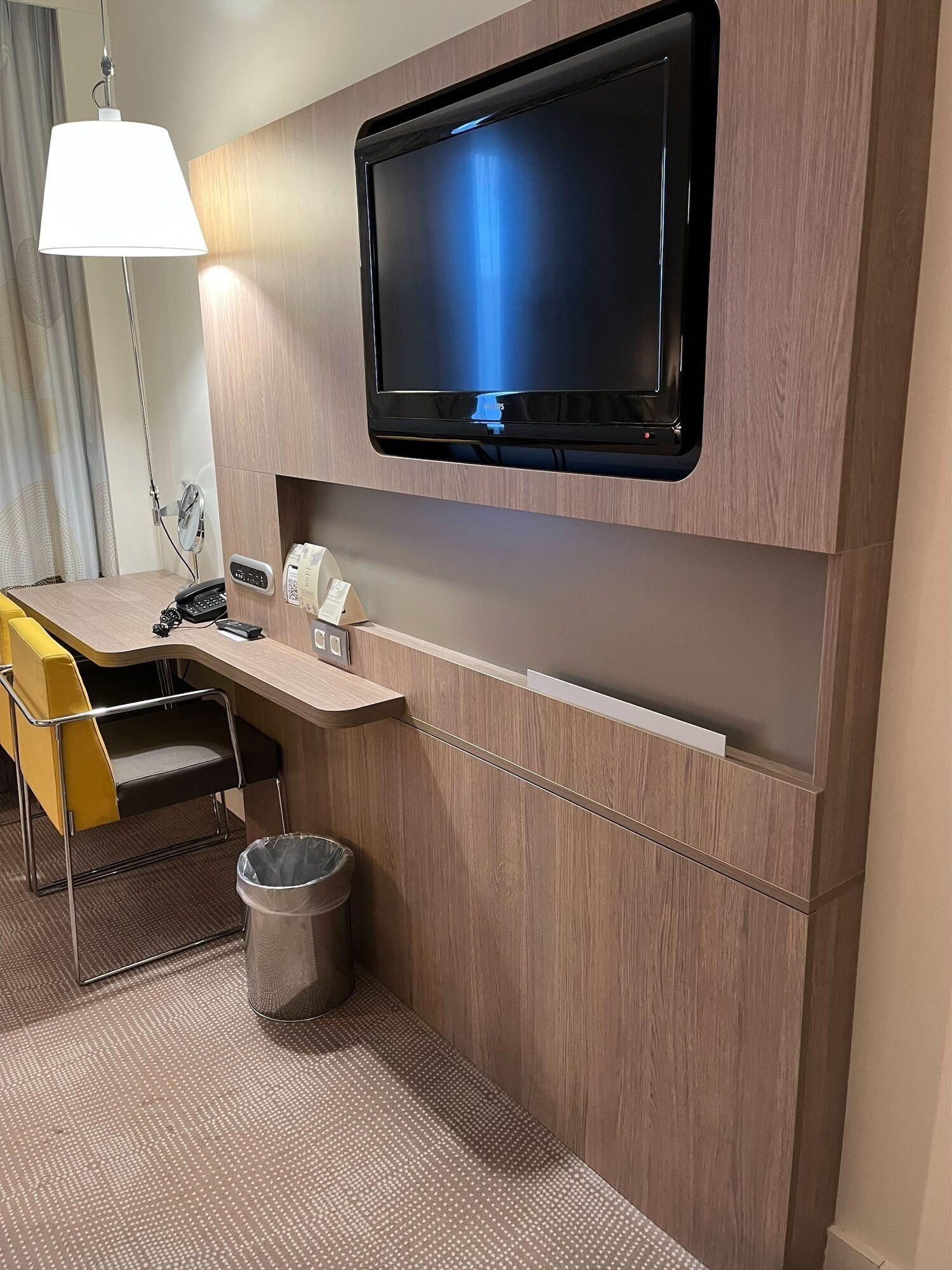 A modern hotel room with a mounted TV, a desk with a chair, a trash bin, and a lamp.