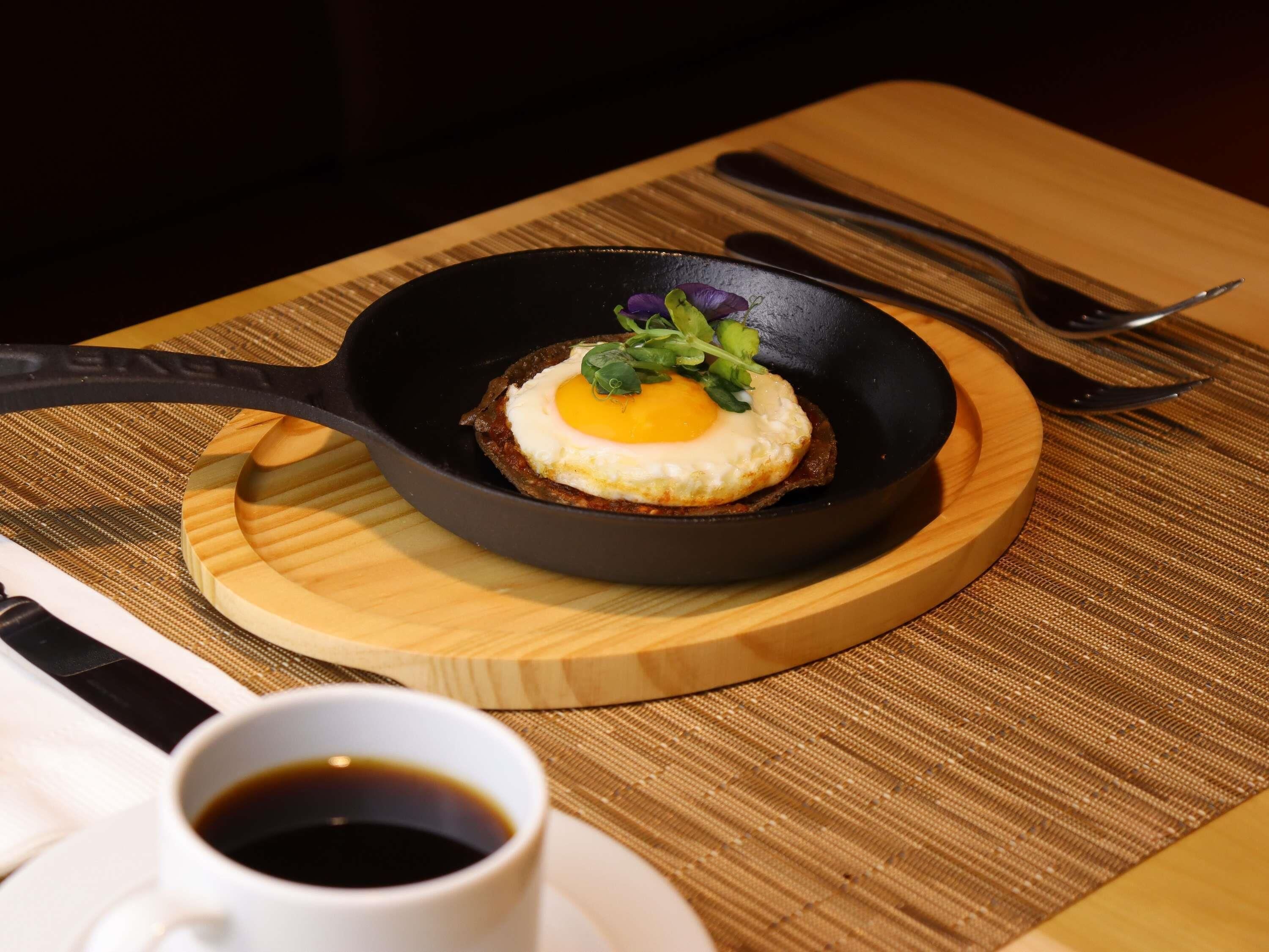 Eine Bratpfanne mit einem Spiegelei auf Toast, eine Kaffeetasse und Besteck auf einem Holztisch.