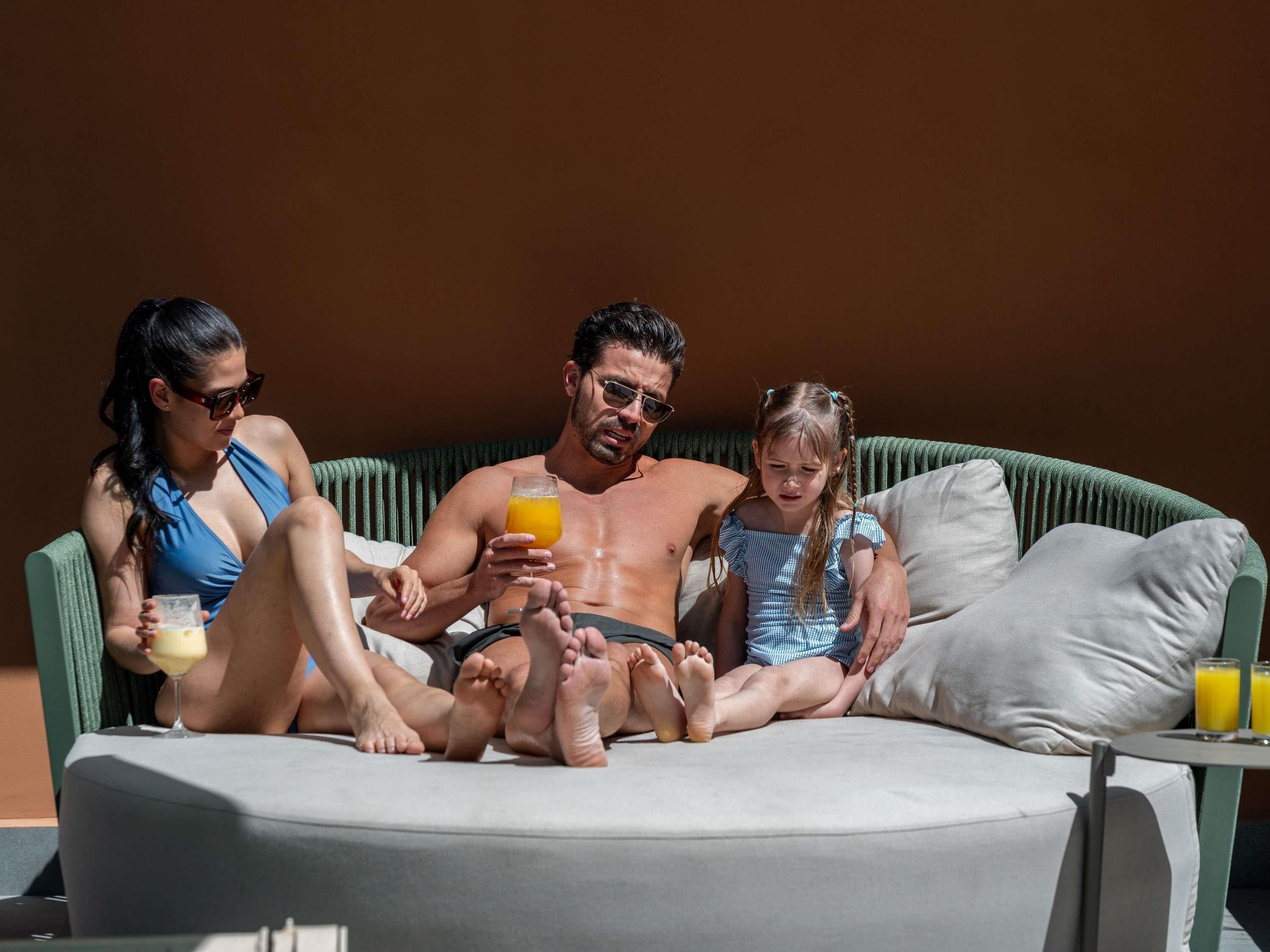 Una familia de tres sentada en un sofá redondo al aire libre, con cojines, una mesita y dos vasos de jugo de naranja.