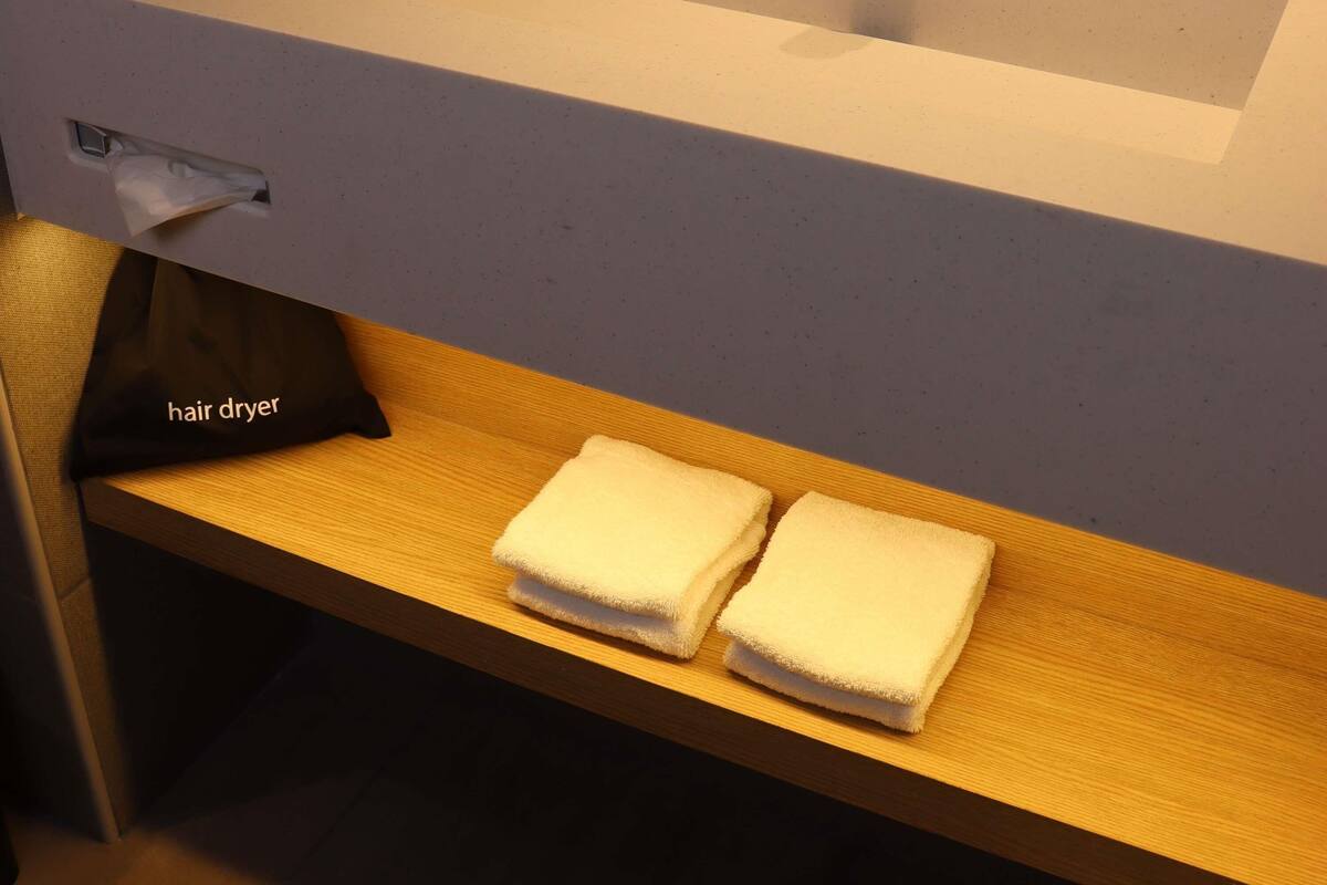 Un baño con un toallero que sostiene dos toallas, un secador de pelo y una bolsa para toallas con la etiqueta 'secador de pelo' sobre un estante de madera.