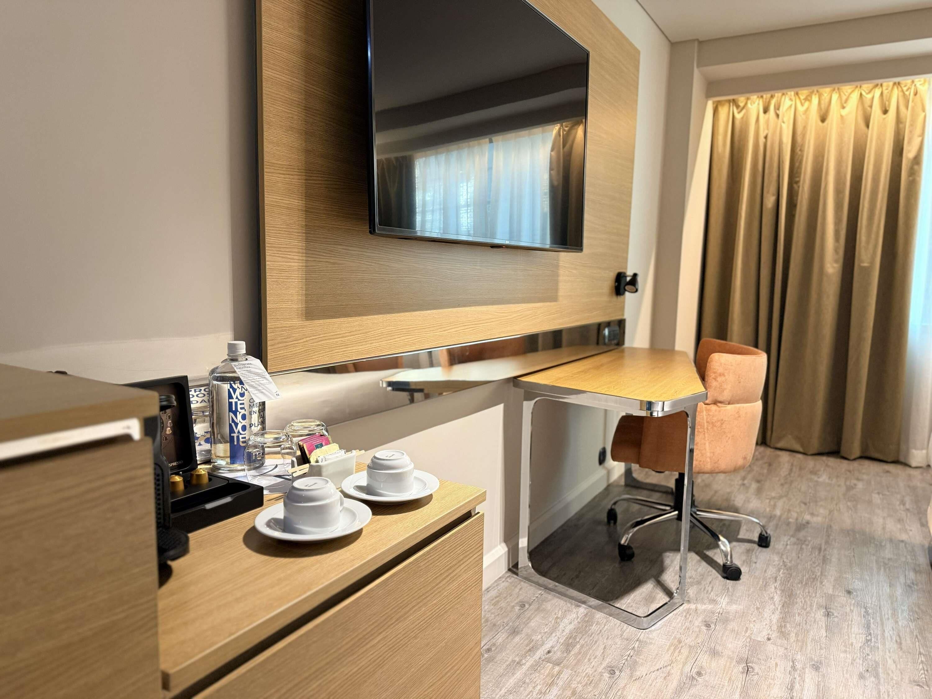 Habitació d'hotel amb escriptori de fusta, televisió penjada a la paret, cadira i finestra amb cortines.