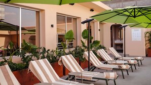 A row of striped lounge chairs under a green umbrella, with potted plants and a beige wall in the background.