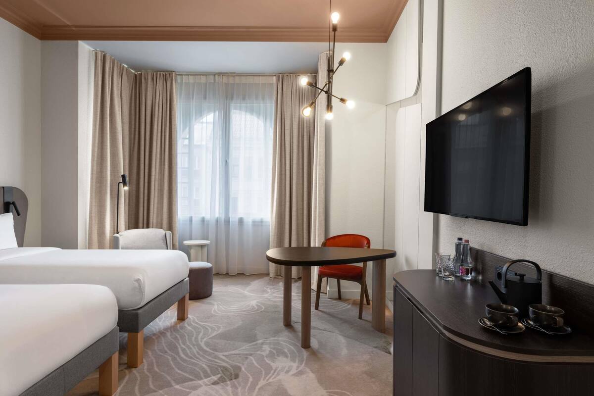 A hotel room with two beds, a TV, a desk with two cups, a chair, and a small table.