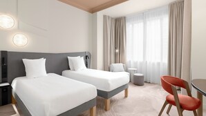 Habitació d'hotel amb dos llits, una taula de treball, una cadira, una làmpada i vista a l'exterior a través de cortines translúcides.