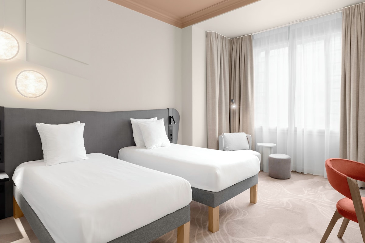 A hotel room with two beds, a desk, a chair, a lamp, and a view of the outdoors through sheer curtains.