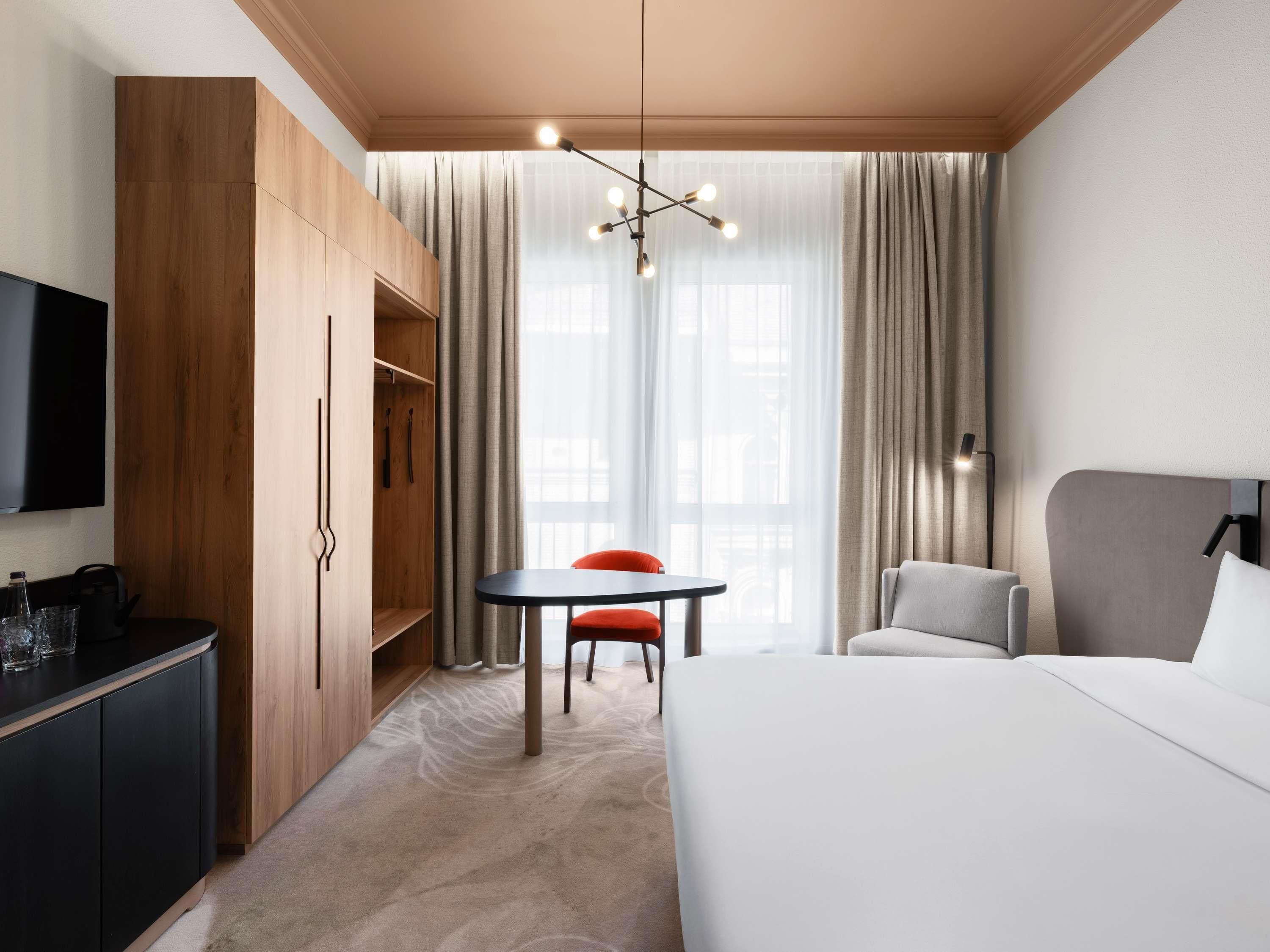 A hotel room with a bed, a desk with a chair, a wardrobe, and a TV.