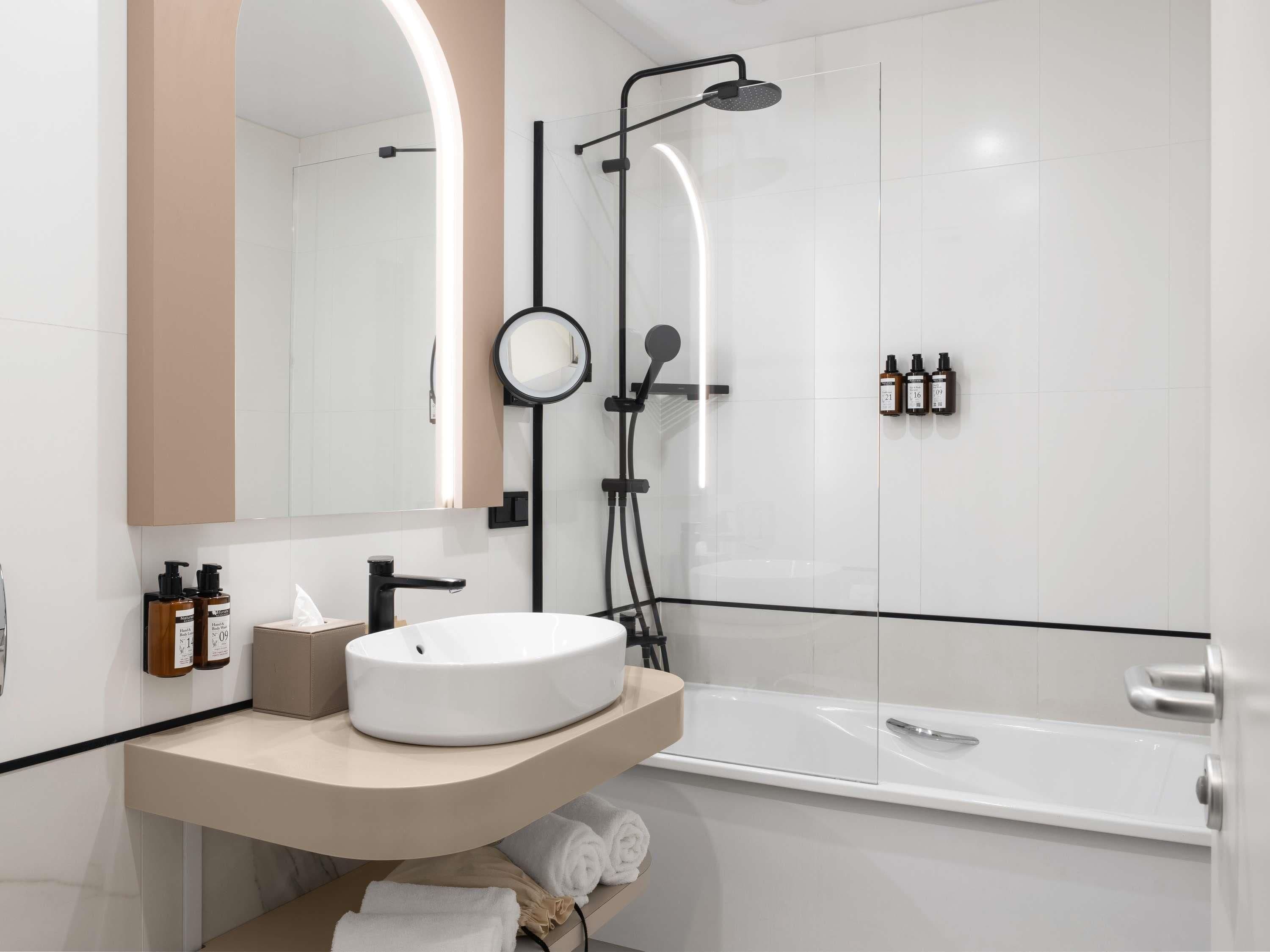 Ein modernes Badezimmer mit einem runden Waschbecken, einer Handbrause, einer Duschkabine aus Glas und einem Spiegel mit rundem Rahmen.