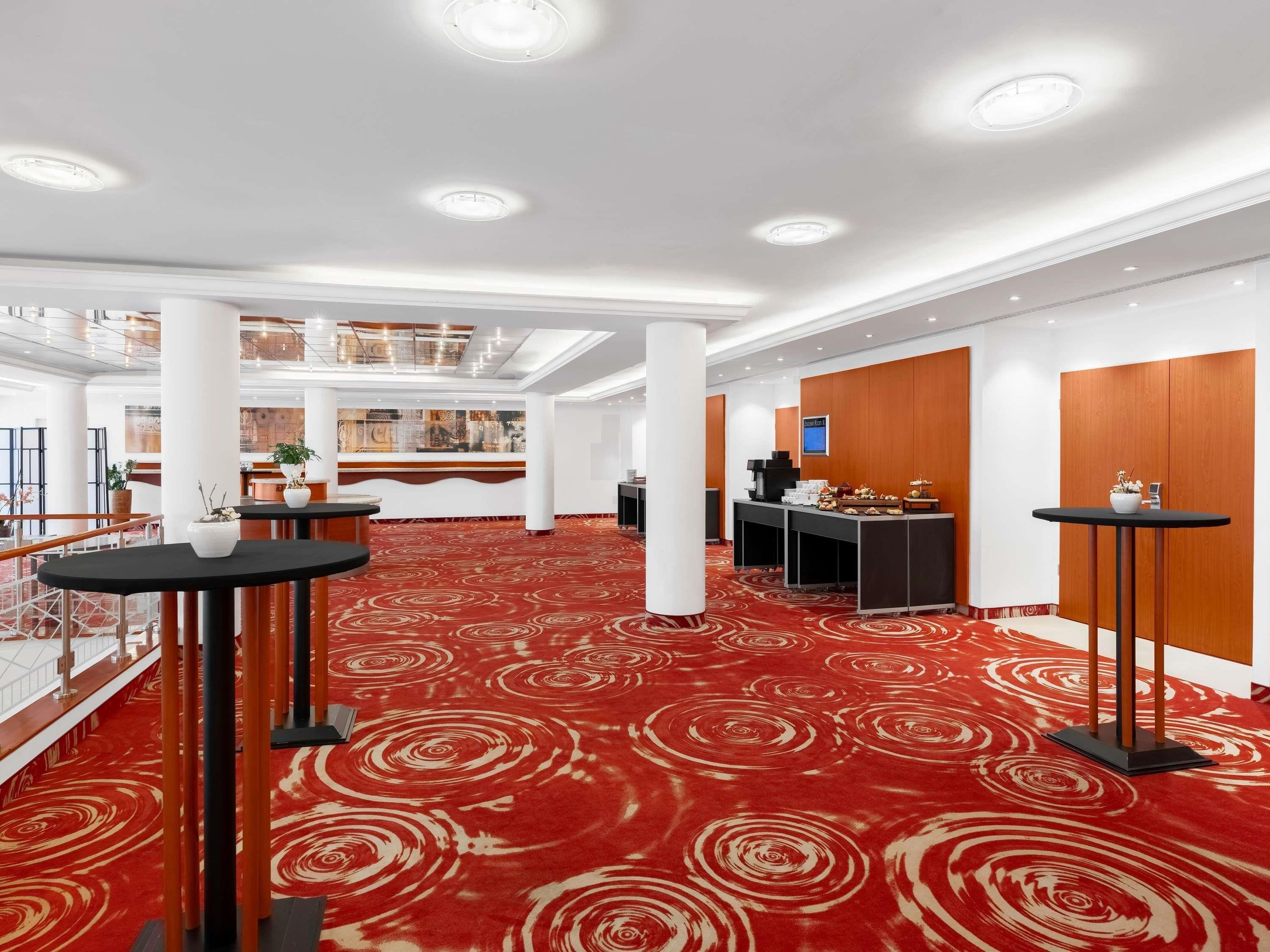A spacious hotel lobby with a red carpet, multiple tables, a reception desk, and a high ceiling with recessed lighting.