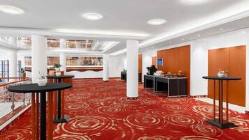 A spacious hotel lobby with a red carpet, multiple tables, a reception desk, and a high ceiling with recessed lighting.