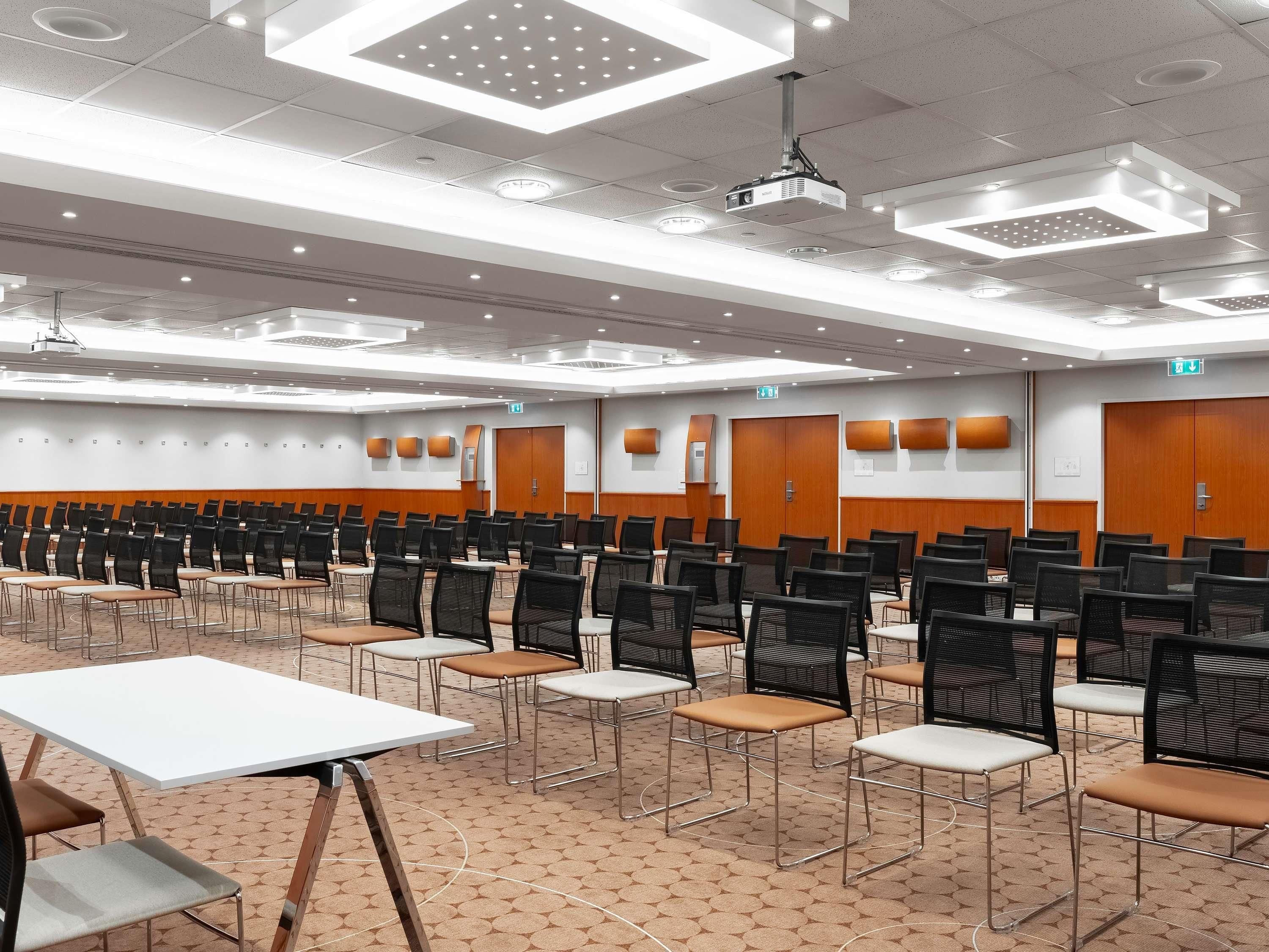 Un salón de conferencias con filas de sillas, un proyector y una mesa blanca.