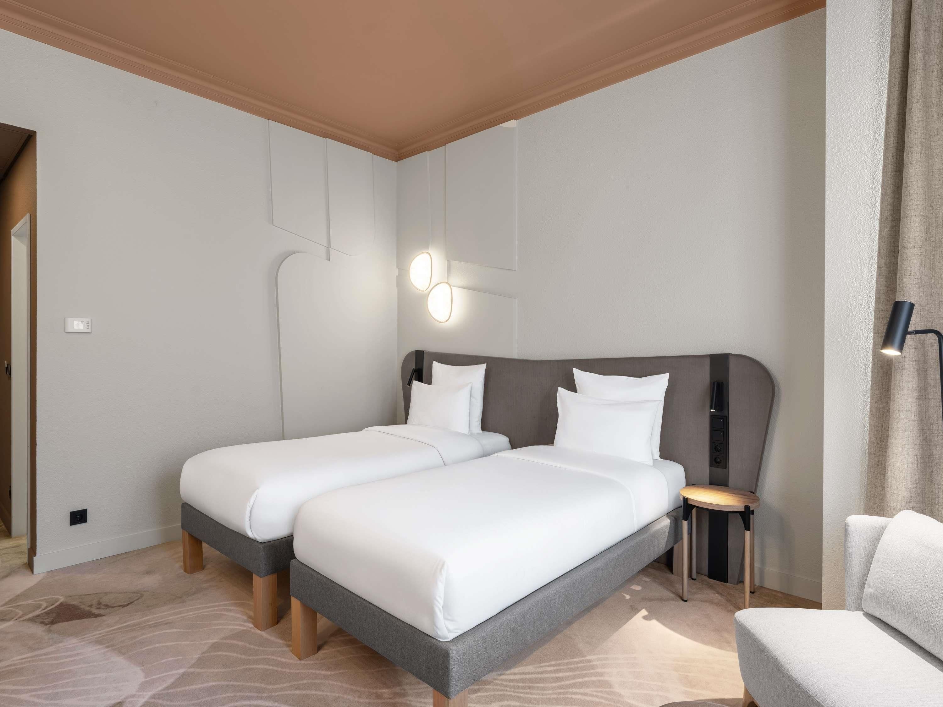 A hotel room with two beds, a small table, a chair, and a lamp.