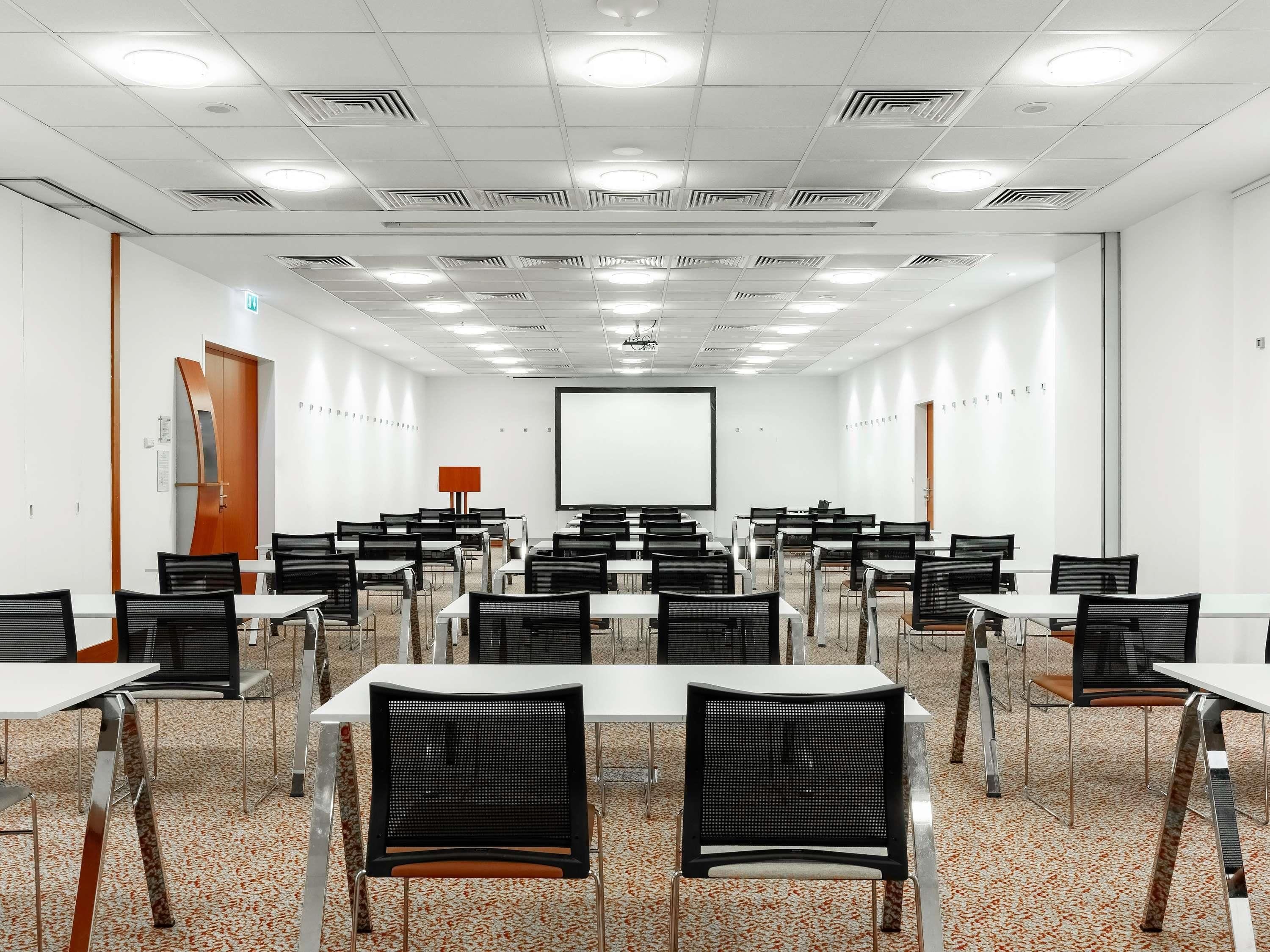 Un salón de conferencias con mesas blancas, sillas negras, una pantalla de proyector y una pizarra.