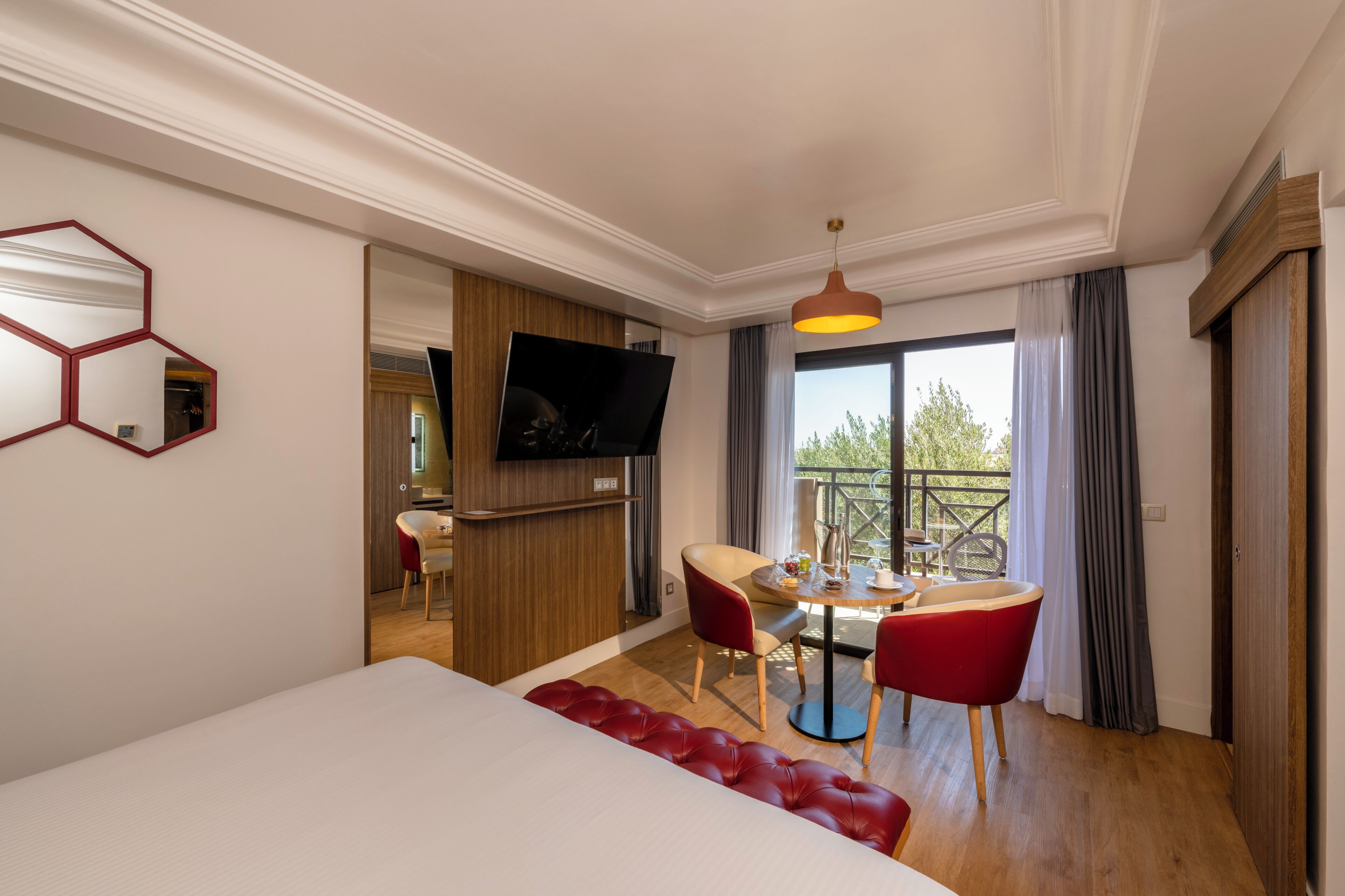 A hotel room with a bed, a dining area with a table and chairs, a TV mounted on the wall, and a balcony with a view.