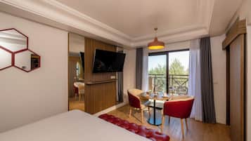 A hotel room with a bed, a dining area with a table and chairs, a TV mounted on the wall, and a balcony with a view.