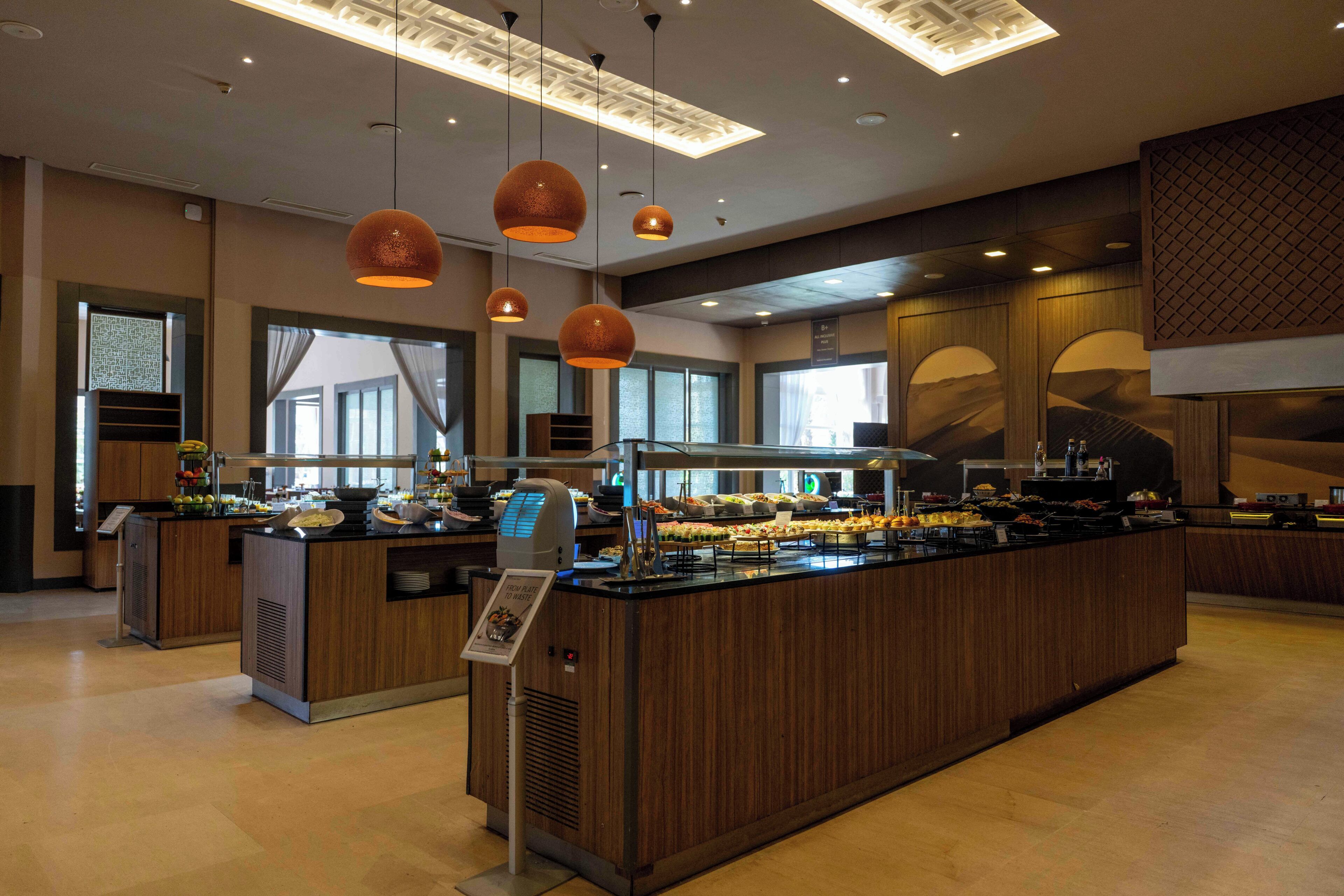 Une cuisine d’hôtel moderne dotée d’un îlot central servant de buffet, d’un éclairage suspendu et de grandes fenêtres.