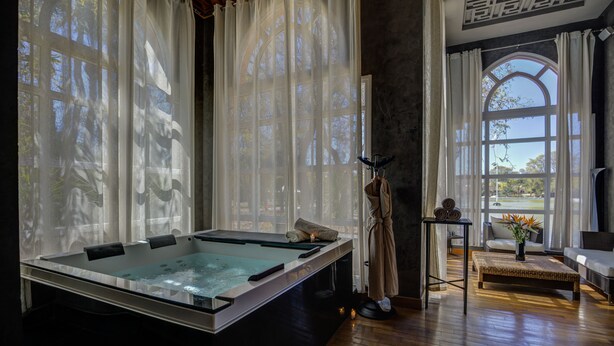 A spa room with a large soaking tub, a massage table, and a view of a garden through large windows.