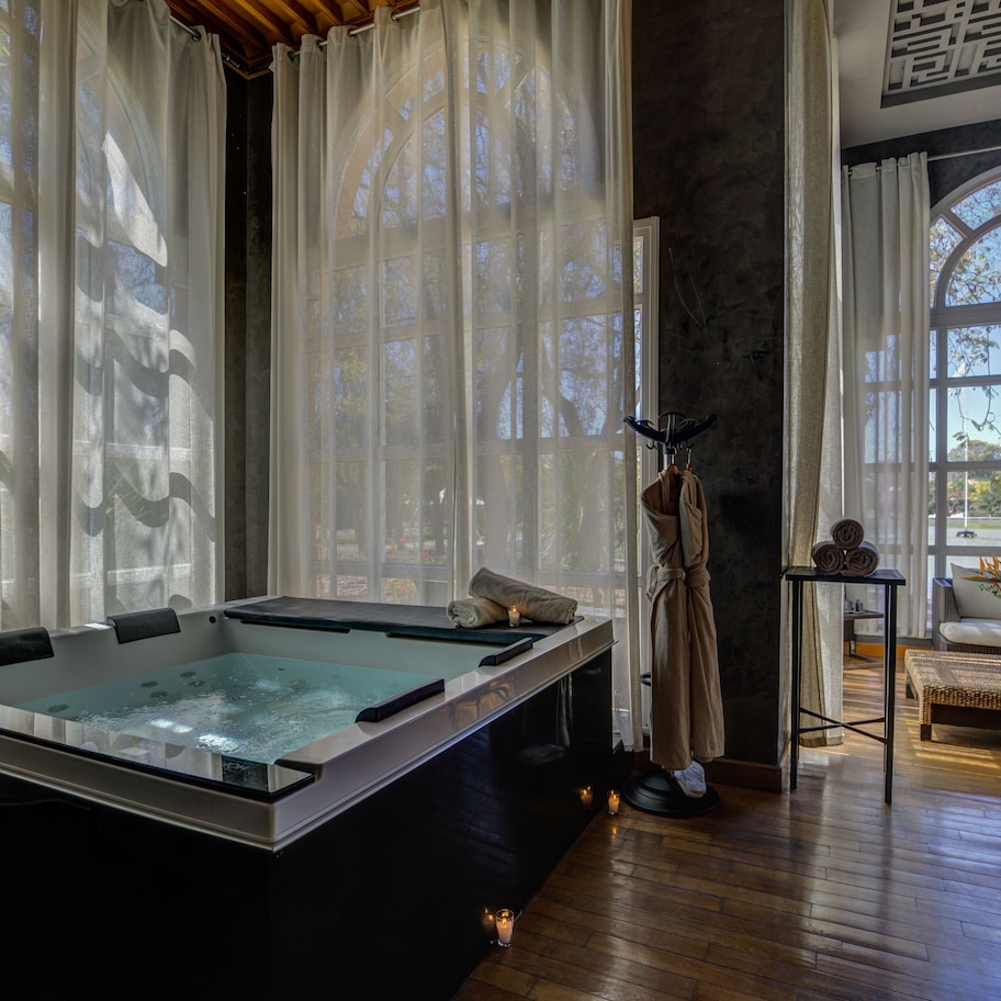 A spa room with a large soaking tub, a massage table, and a view of a garden through large windows.