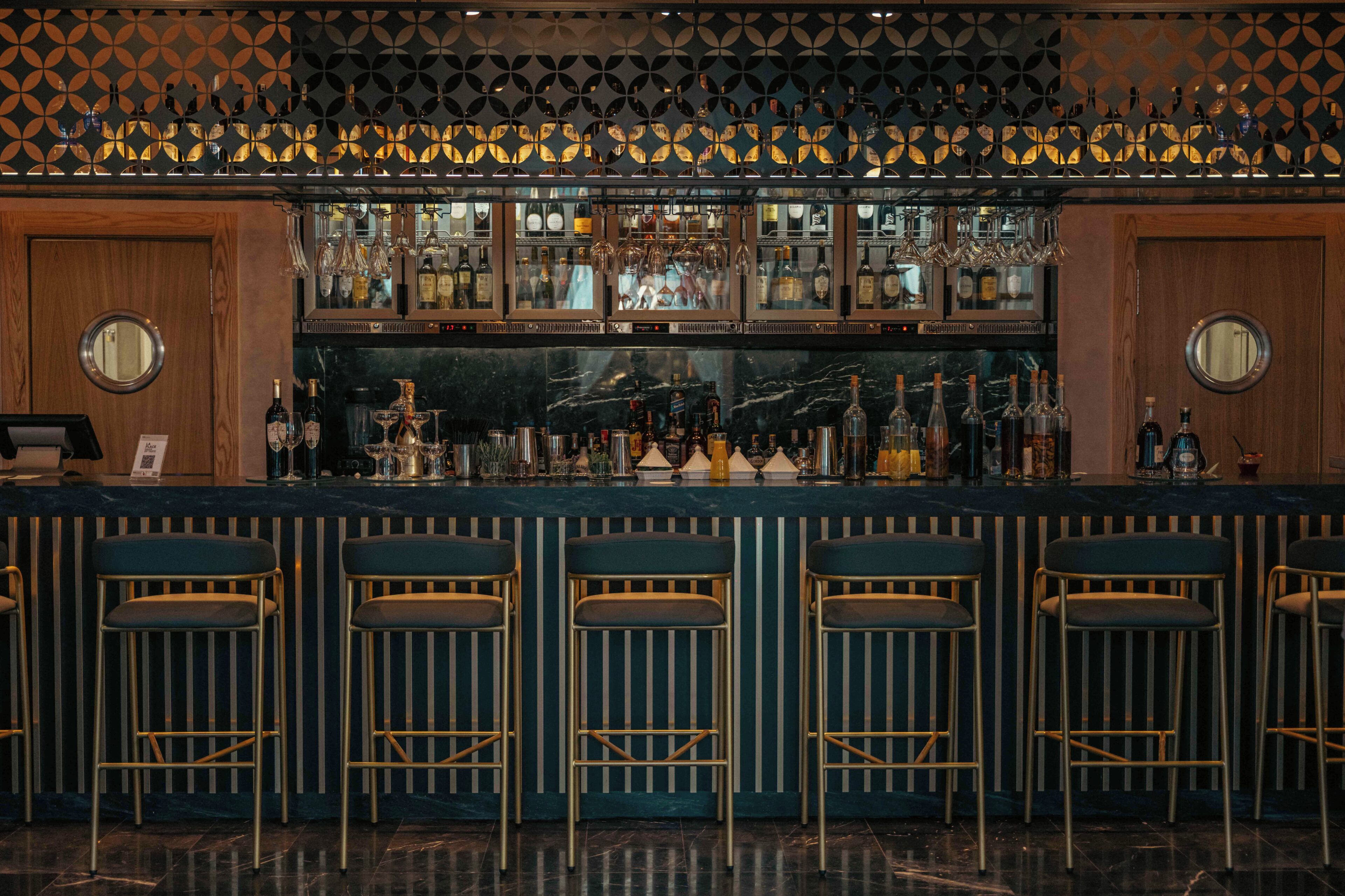 Un bar avec un décor mural à motifs, de nombreuses étagères remplies de bouteilles et des tabourets de bar.