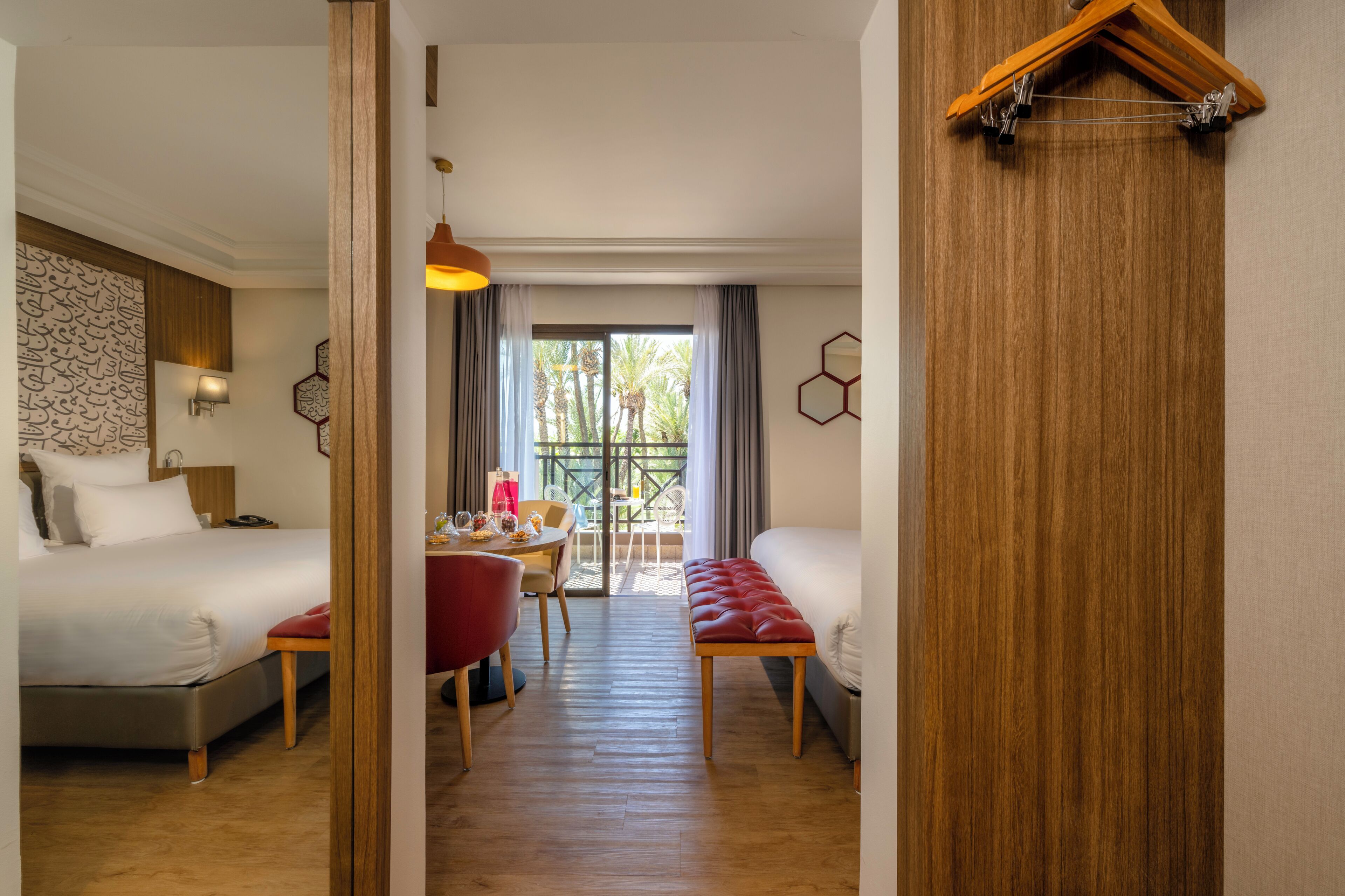 A hotel room with two beds, a dining table, and a view of palm trees through the window.
