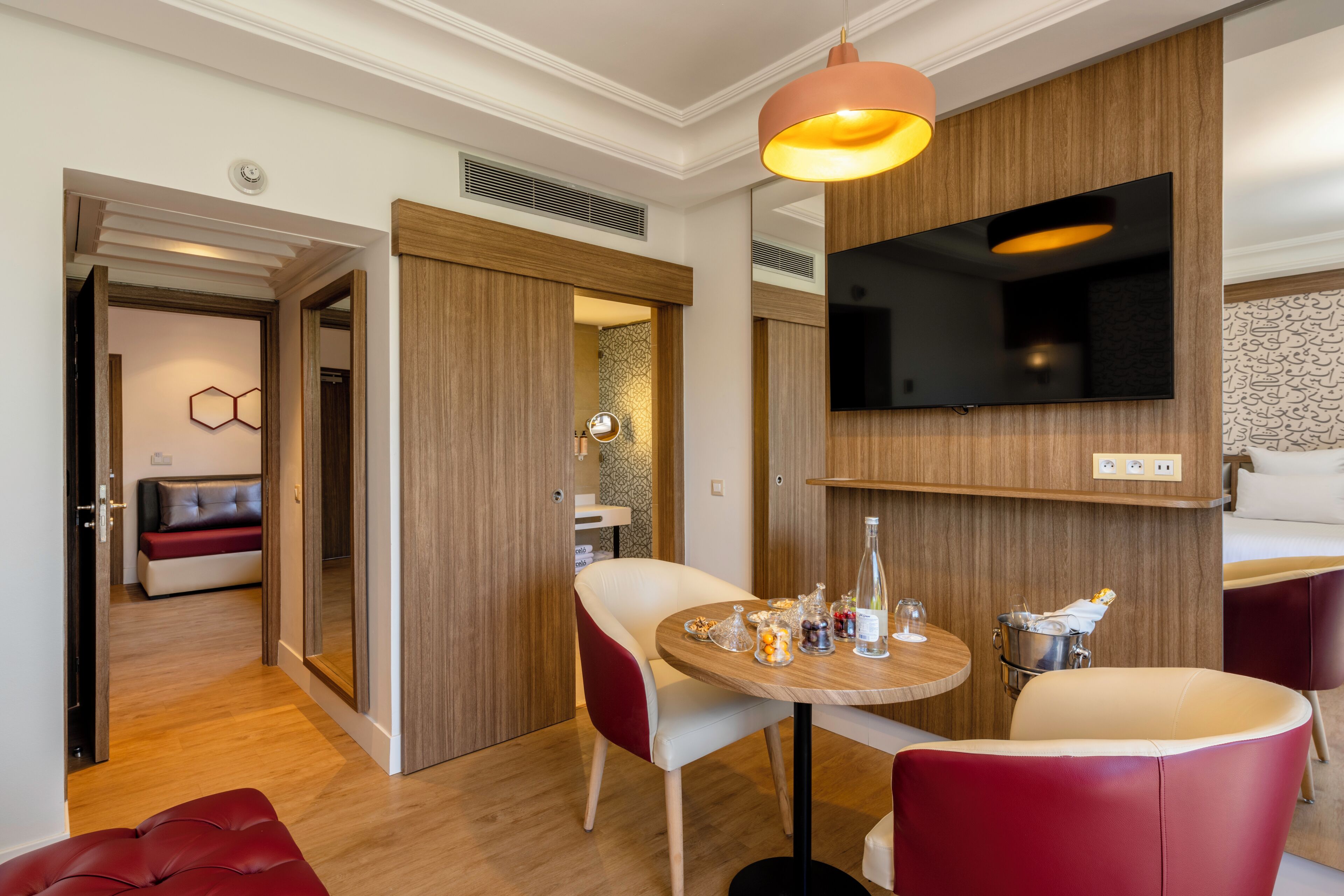 A modern hotel room with a wooden wall panel, a flat-screen TV, a round wooden table with a bottle of wine, and a red leather sofa.